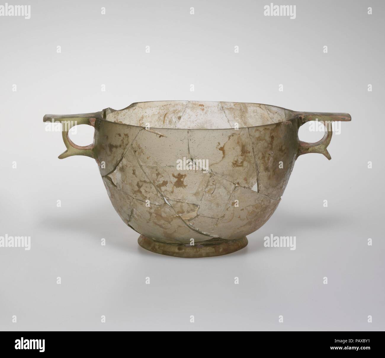 Glas skyphos (Trinkbecher). Kultur: Griechisch. Abmessungen: H.: 5 7/8 in. (14,9 cm); Durchm. (Rim): 8 3/8 in. (21,3 cm). Datum: Ende 3./Anfang 2.Jh. v. Chr.. Farblos mit hellen grünliche Färbung. Leicht invers Rim, mit der Oberkante Boden flach; konvex geschwungene Seite nach unten spitz zulaufend; gespreizter Fuß Ring mit abgerundeten unteren Rand; leicht hügelig, flacher Boden, zwei Griffe an den Seiten des Körpers angewandt, die von Leerzeichen durch erhöhte Fläche umgeben geschnitzt, mit Flachbild Daumen - liegt Ringe und projizieren Flügel oben und unten. Körper gebrochen und repariert, mit einem großen Chip in Rim und Sev Stockfoto
