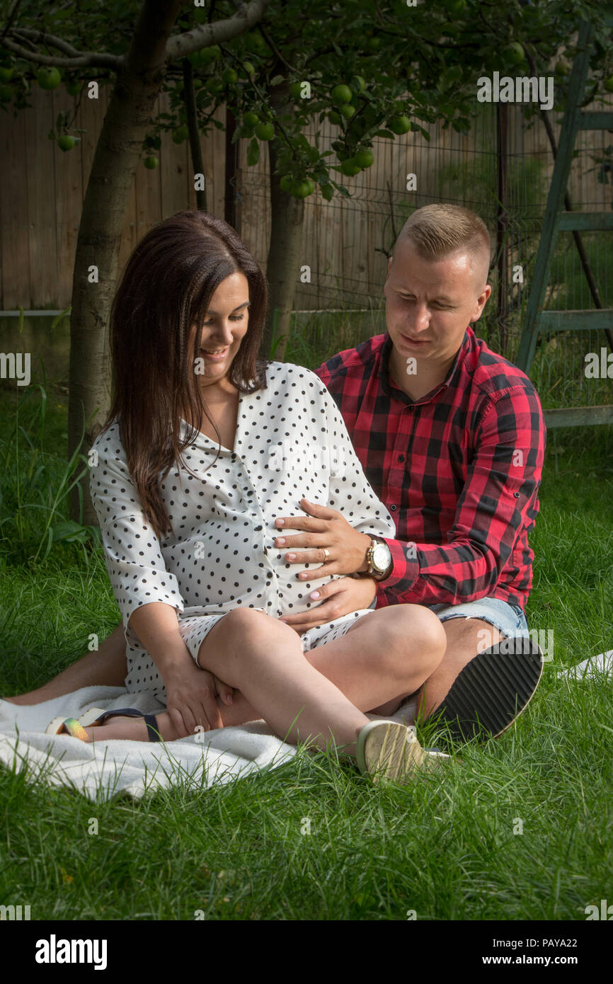 Junge schwangere Frau und ihr Mann im Garten sitzen und beobachtete, wie die Bewegungen des Kindes noch geboren werden. Stockfoto