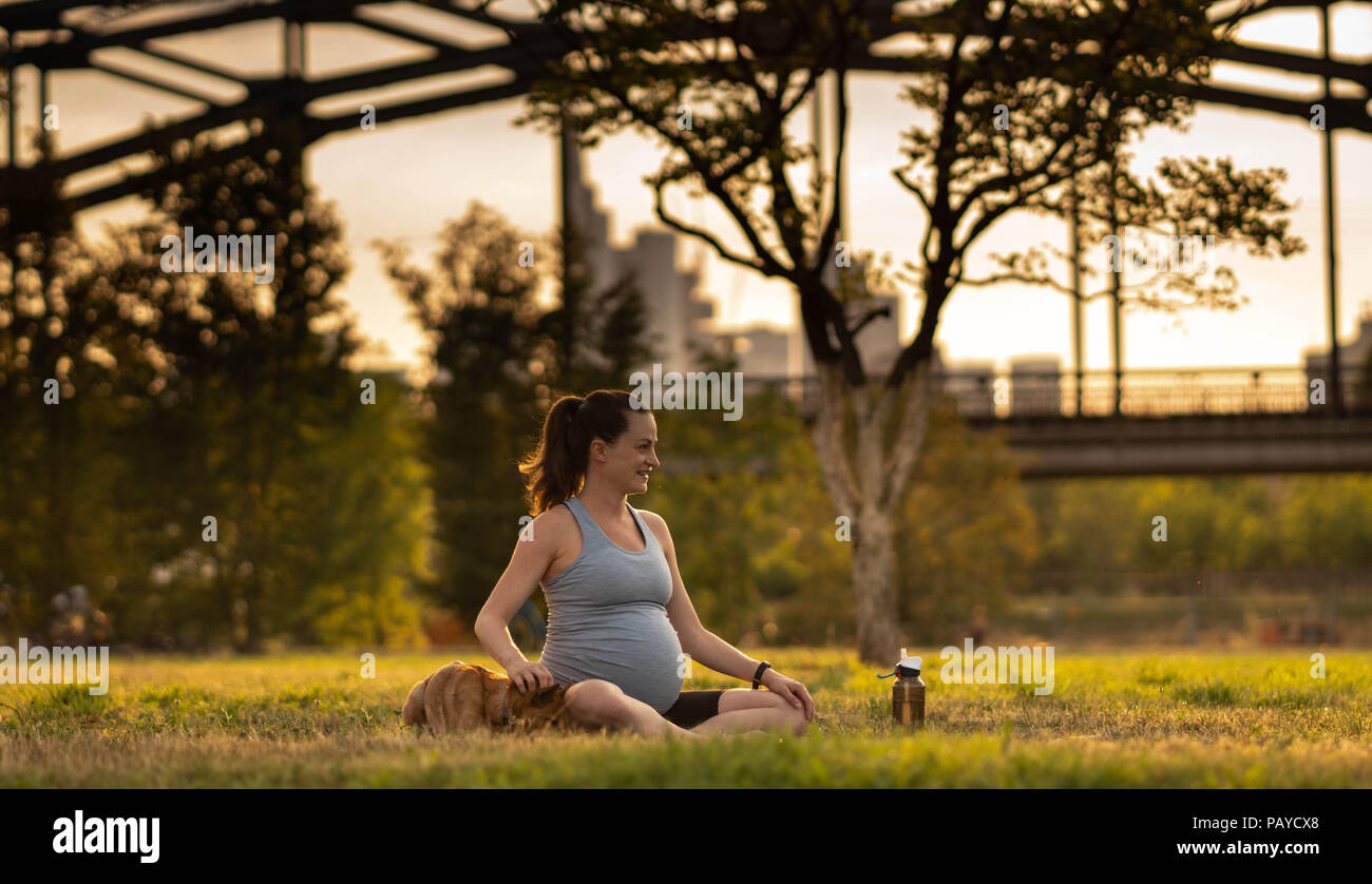 Schöne schwangere junge Frau mit kleinen Hund sitzen auf Matten, Yoga, Entspannung, Gefühl, lebendig, frische Luft, Ruhe und Träumen, im grünen Park. Auf dem Hintergrund der Eisenbahnbrücke und der Stadt Stockfoto