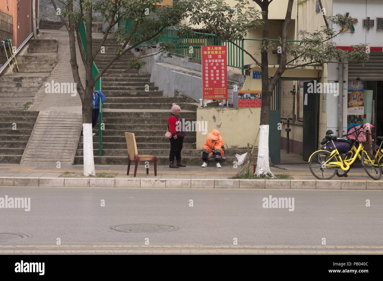 Kunming, Yunnan, China - 30 Dezember 2017: Zwei chinesische Frauen auf dem Bürgersteig mit einem Stuhl Stockfoto