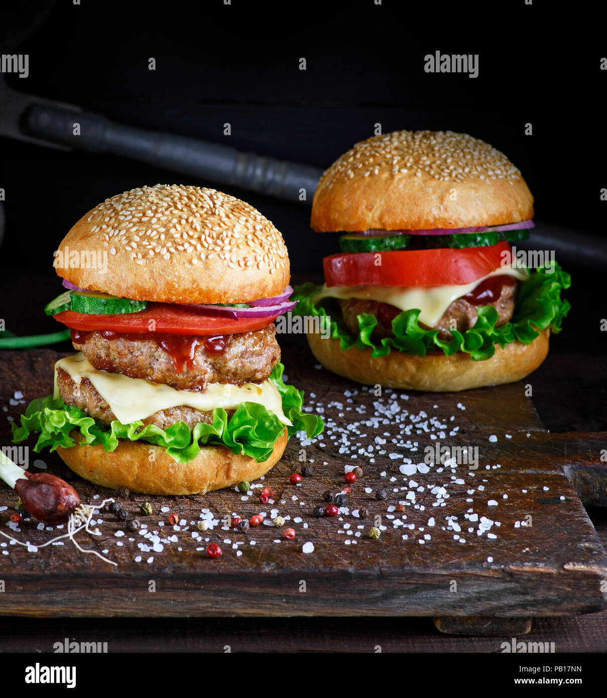 Zwei Burger mit Fleisch hacken, Gemüse und grünen Salat in eine runde Brötchen Mit Sesam auf braunem Holz- Board, Gewürze sind in der näheren Umgebung verstreut Stockfoto