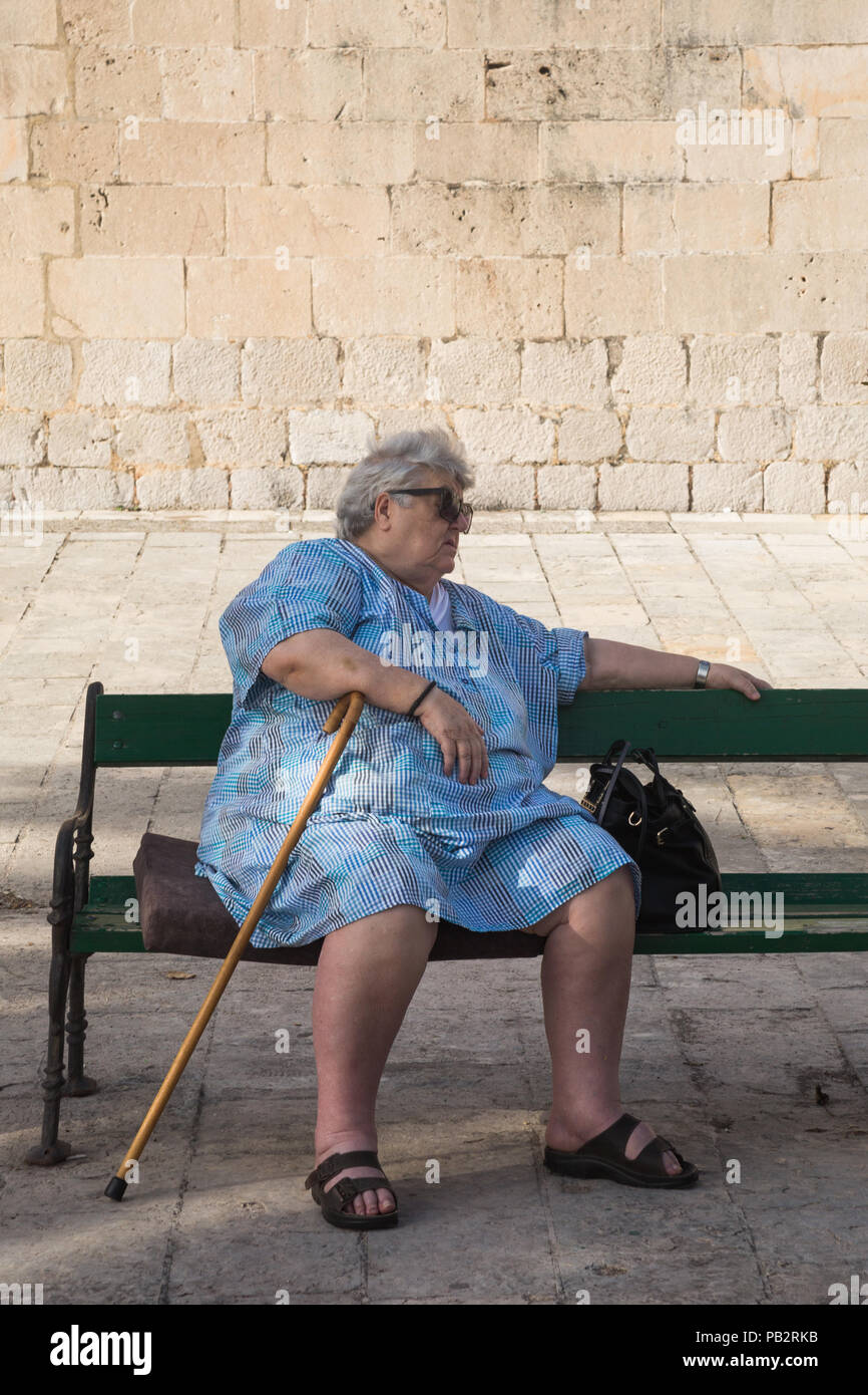 Ältere Frau im blauen Kleid mit Stock und Sonnenbrille setzte sich auf grünen Holz- Sitzbank in Schatten außerhalb St. Nikola Kirche, Perast, Montenegro. Stockfoto
