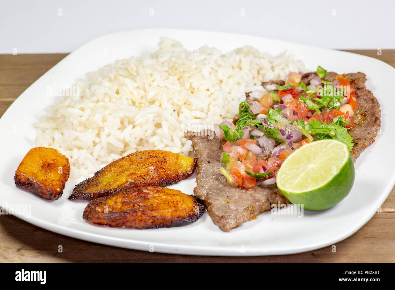 Ein Steak in Pico de Gallo von Kochbananen und weißen Reis auf einem weißen Teller umgeben. Kubanisches Essen. Stockfoto