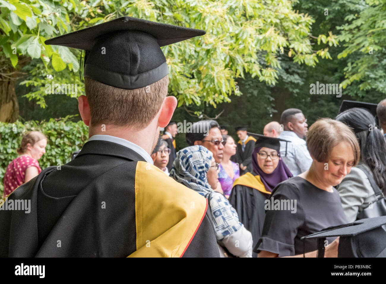 Junge männliche Graduierten Student sah seine Graduierung Kleid an einem Campus Graduierung Party zeigte verschiedene Eltern im Hintergrund. Stockfoto