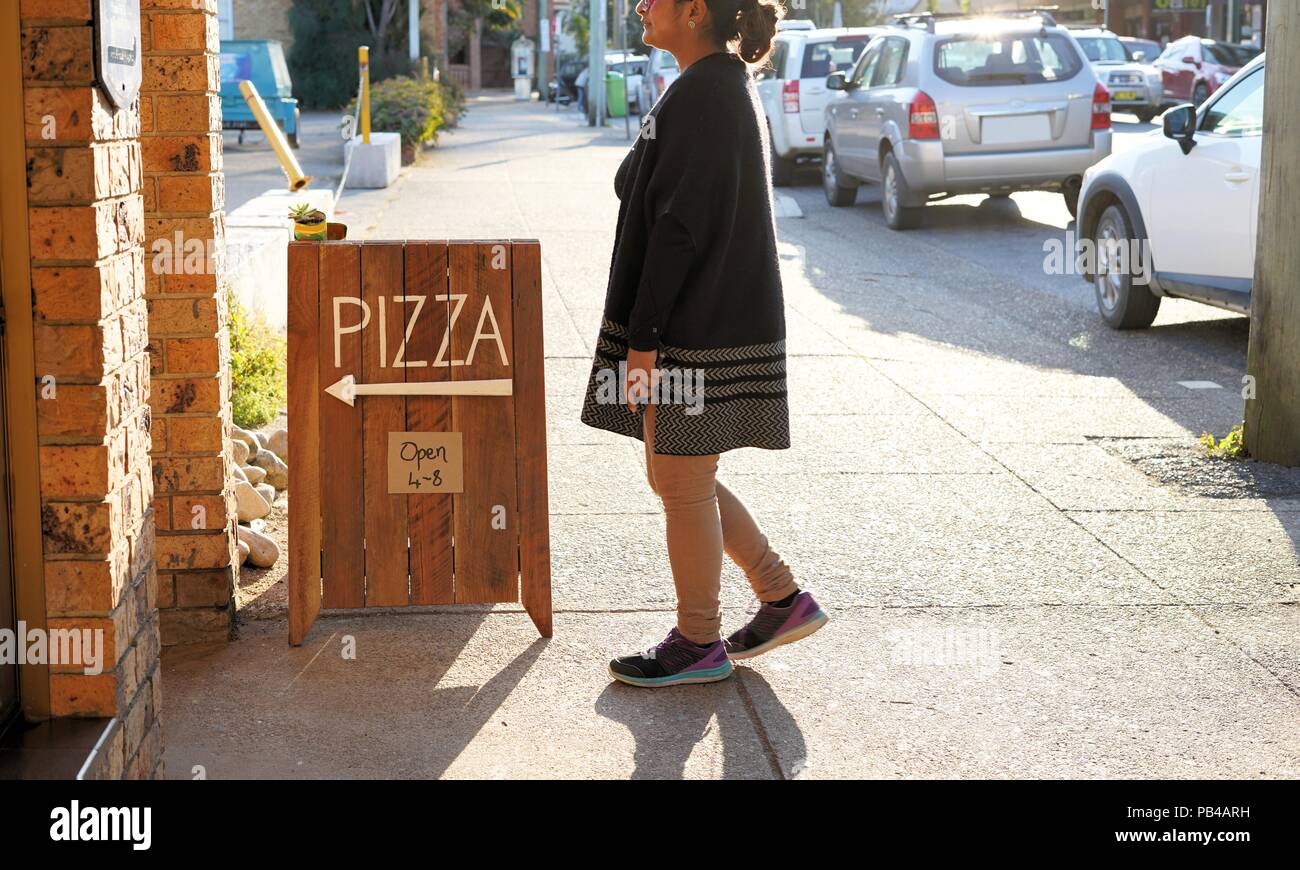 Frau wandern in Pizza Restaurant. Ehrliches Bild der Frau vor der Pizza Schild außerhalb Pizza Shop in der Nähe der Straße mit Autos geparkt. Stockfoto