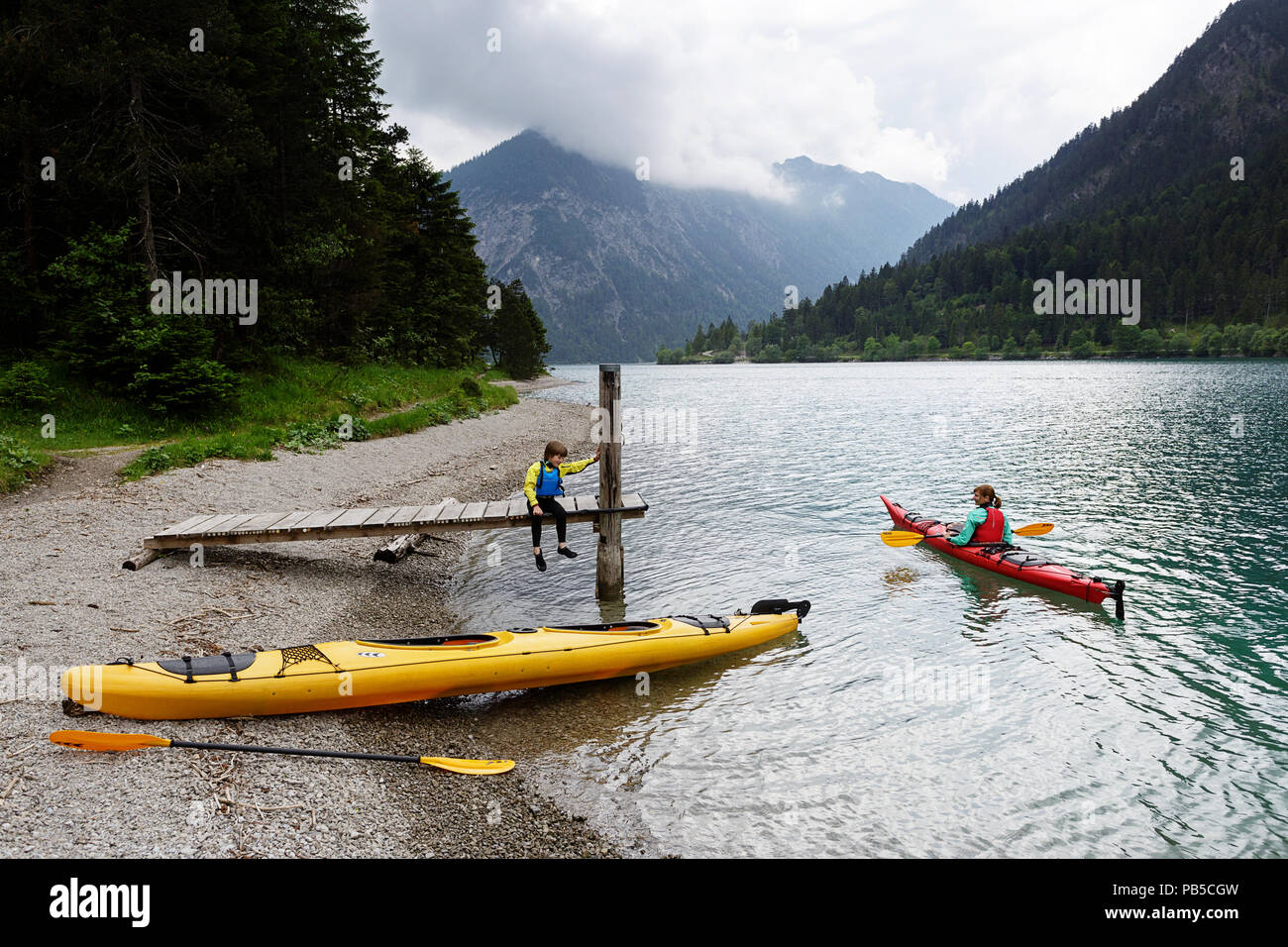 Mutter in einem Kajak im Gespräch mit ihrem Sohn sitzen auf dem hölzernen Pier mit einem Kajak am Ufer am Plansee, Österreich geparkt Stockfoto