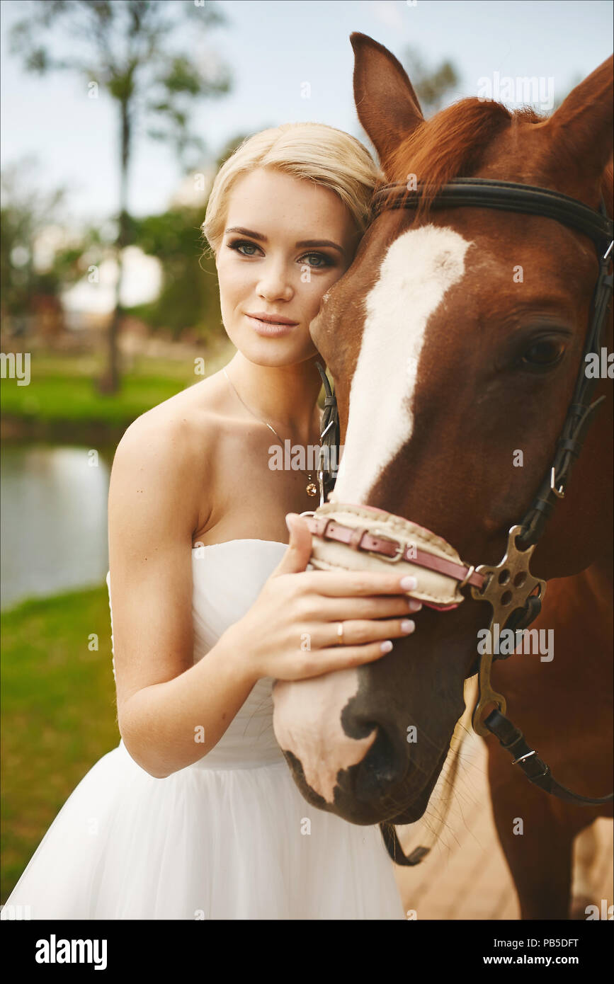 Schöne und modische junge Braut, blonden Model Mädchen mit blauen Augen und  stilvolle Frisur in weißem Kleid mit braunen Pferd im Freien posieren  Stockfotografie - Alamy