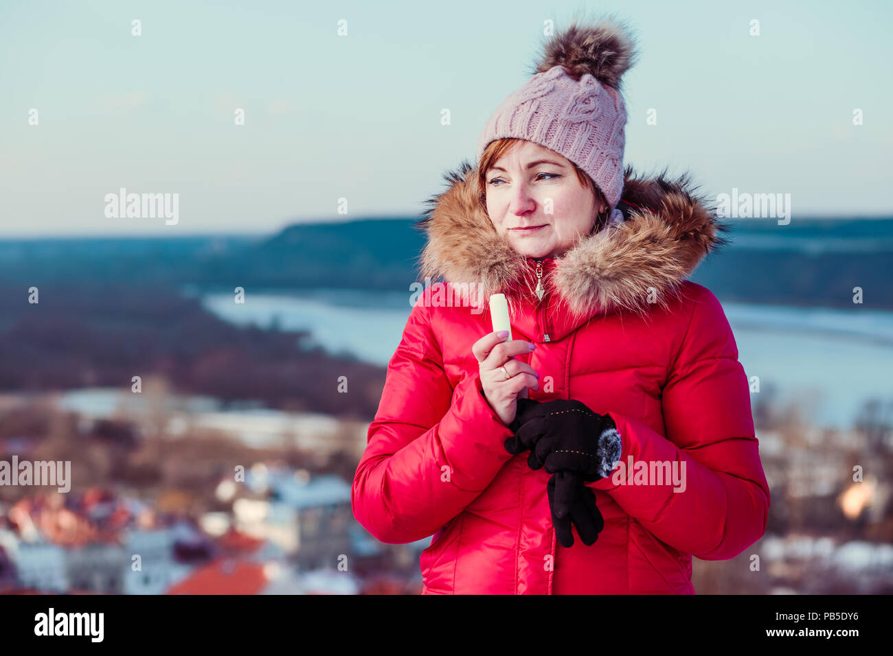 Frau Anwendung Lippenbalsam beim Spaziergang in einem Wintertag. Das Tragen der roten Mantel und Mütze. Stadt im Hintergrund Stockfoto