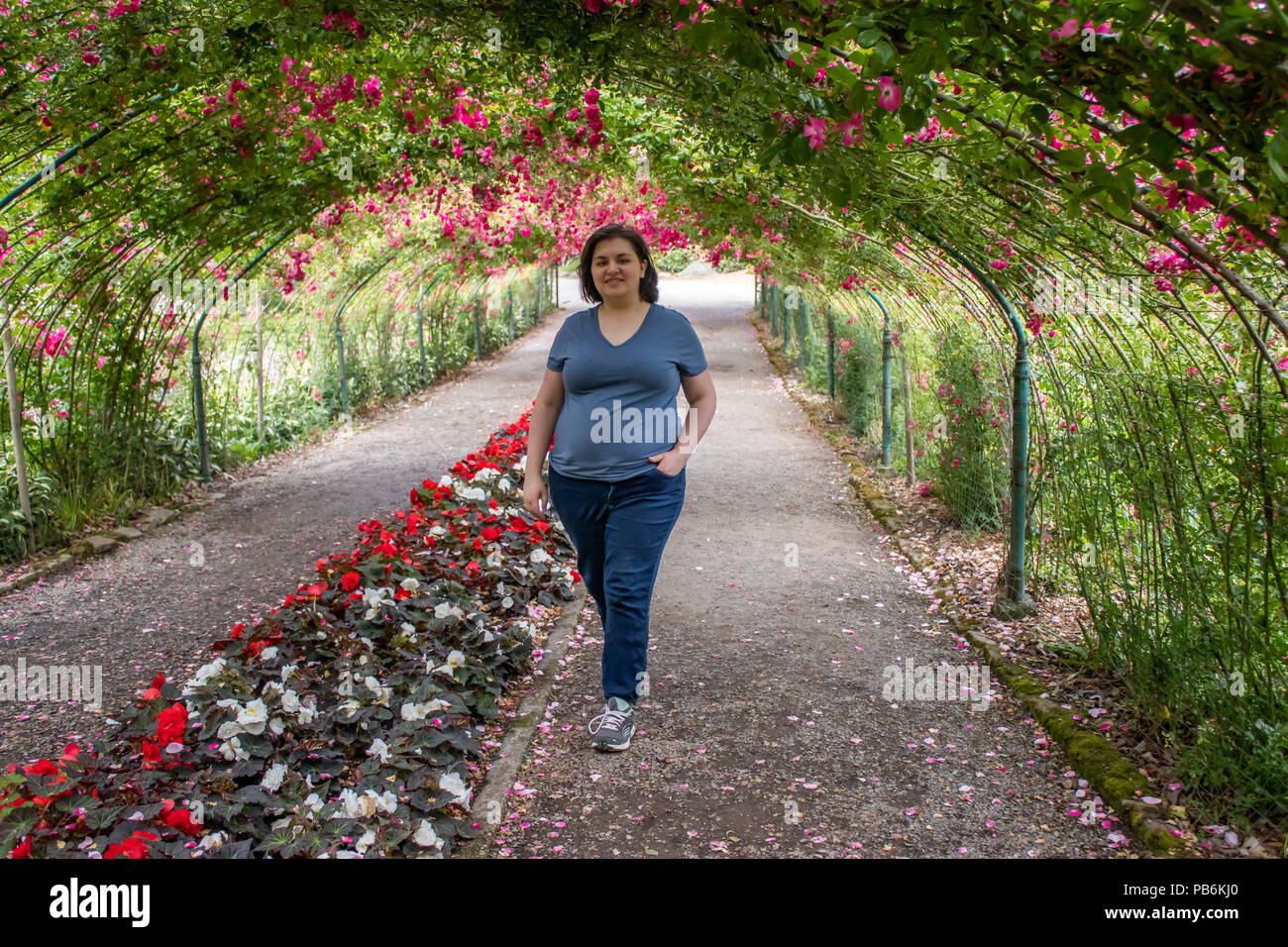 Junge Erwachsene brünette Frau in einem blauen Hemd mit kurzen Haaren zu Fuß durch einen Rosengarten tunnel Stockfoto