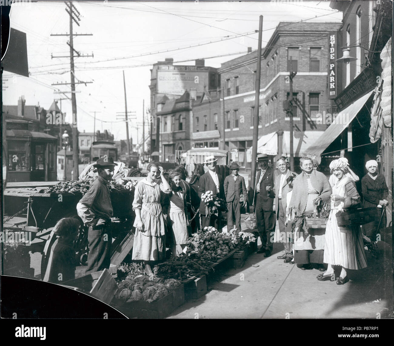 . Englisch: Horizontal, Schwarz und Weiß Foto, Käufer von bürgersteig produzieren außerhalb des Theodore Prieshoff Lebensmittelgeschäft an der East Grand Avenue 1929 versammelt, in der Nähe der Grand Avenue Wasserturm. Das Produkt ist auf einer Plattform auf dem Bürgersteig arrangiert, und eine kleine Gruppe von Männern und Frauen ist um gesammelt. Einige Käufer halten Körbe voll produzieren. Mehrere Autos und ein Pferd und Wagen kann auf der linken und die Theodore Prieshoff Lebensmittelgeschäft können auf der rechten Seite gesehen werden gesehen werden. Eine Vielzahl von 2- und 3-stöckige Gebäude im Hintergrund. Das Drucken wird angezeigt wurden her zu haben Stockfoto