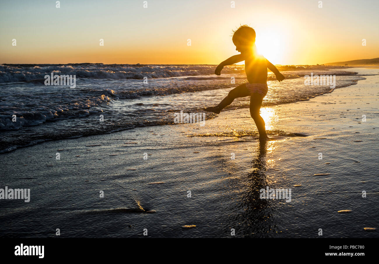 Baby Boy Spielen macht Spritzwasser bei Sonnenuntergang. Spanisch Südwesten Strand. Er ist das Treten der Wellen Stockfoto