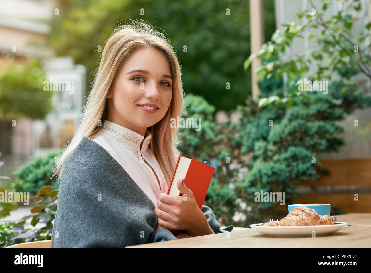 Cute girl Holding Big Red Book Cafe im Freien sitzen, essen leckere Croissants. In kurze blonde Haare, große blaue Augen, blassen Wangen, dicke Lippen. Grüne Pflanzen und Sommer Straße für den Hintergrund. Stockfoto