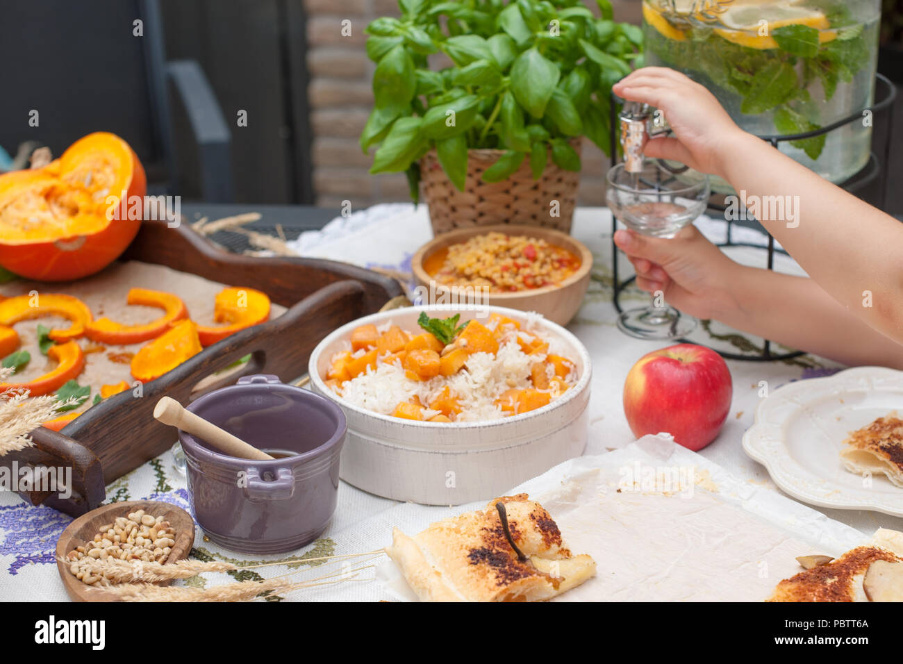 Reis mit Kürbis und Honig auf dem Tisch und andere Herbstspeisen, Kuchen mit Birne. Köstliches Familienessen im Innenhof. Baby Hände gießen Limonade. Stockfoto