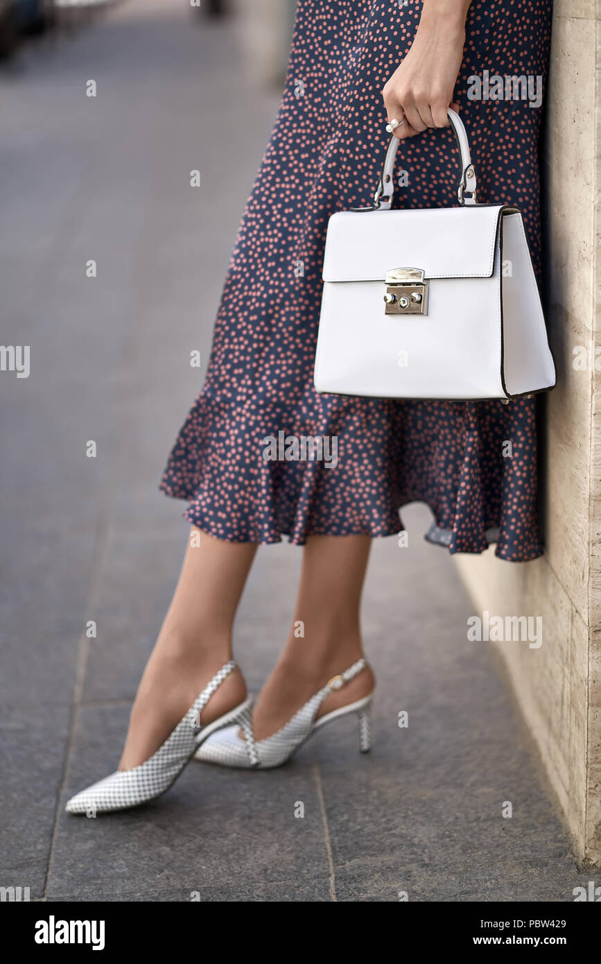 Schöne Mädchen posiert mit einem weißen Beutel auf der Wand Hintergrund im Freien. Sie trägt ein bunt gepunkteten Kleid und Schuhe. Closeup vertikale Foto. Stockfoto