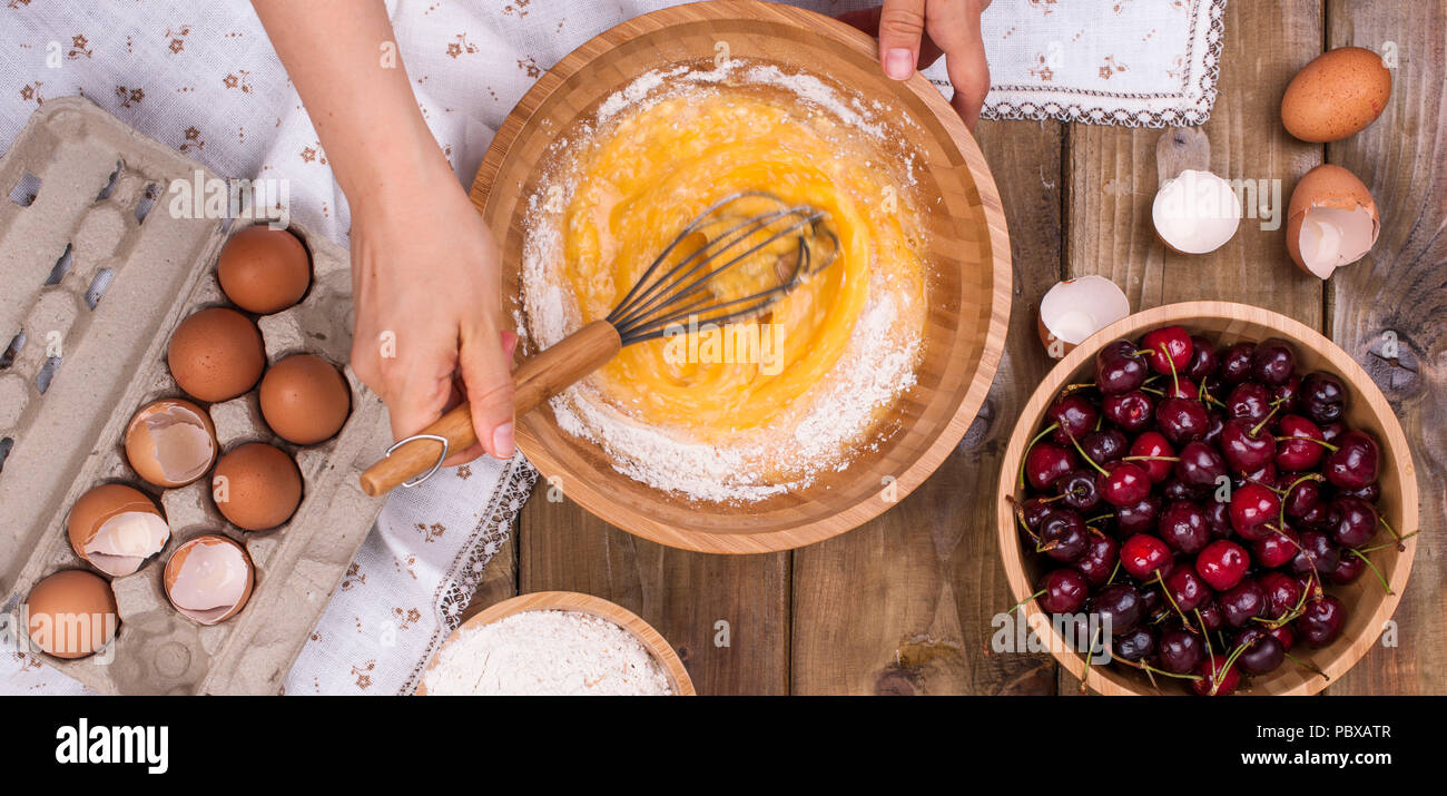 Zubereitung von hausgemachtem Süßgebäck mit Kirschen. Eier, Mehl und Beeren auf Holzgrund. Hände in den Rahmen. Flaches Lay, Banner. Stockfoto