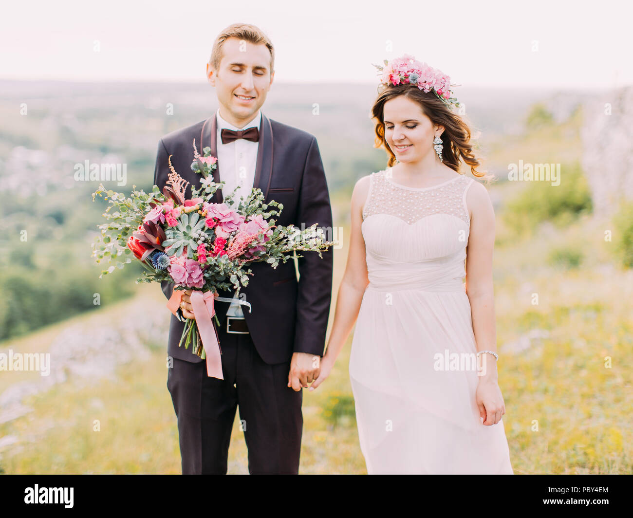 Der lächelnde jungvermählte Paar mit geschlossen Augen halten sich an den Händen. Der Bräutigam hält den Brautstrauß. Die horizontale Nahaufnahme. Stockfoto