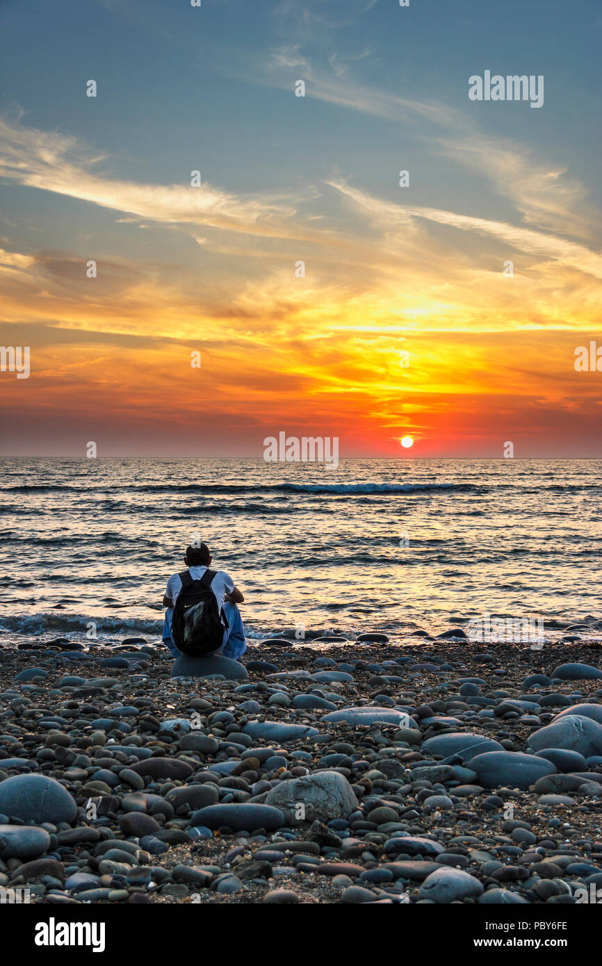 Ein Mann sitzt auf einem steinigen Strand beobachten die dramatischen Sonnenuntergang am Westward Ho!, Devon, Großbritannien Stockfoto
