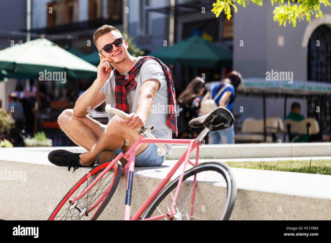 Hübscher junger Mann in hipster Sonnenbrille in der Nähe der Fahrrad sitzen, lächeln und sprechen auf dem Smartphone im sonnigen Straße Stockfoto