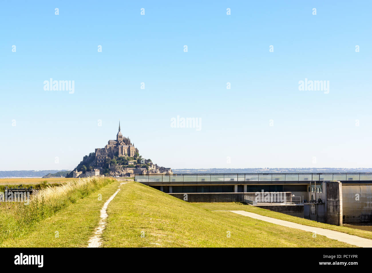 Blick auf den Mont Saint-Michel tidal Island in der Normandie, Frankreich, mit dem Damm auf der Couesnon river und ein Weg entlang des Flusses in den Vordergrund. Stockfoto