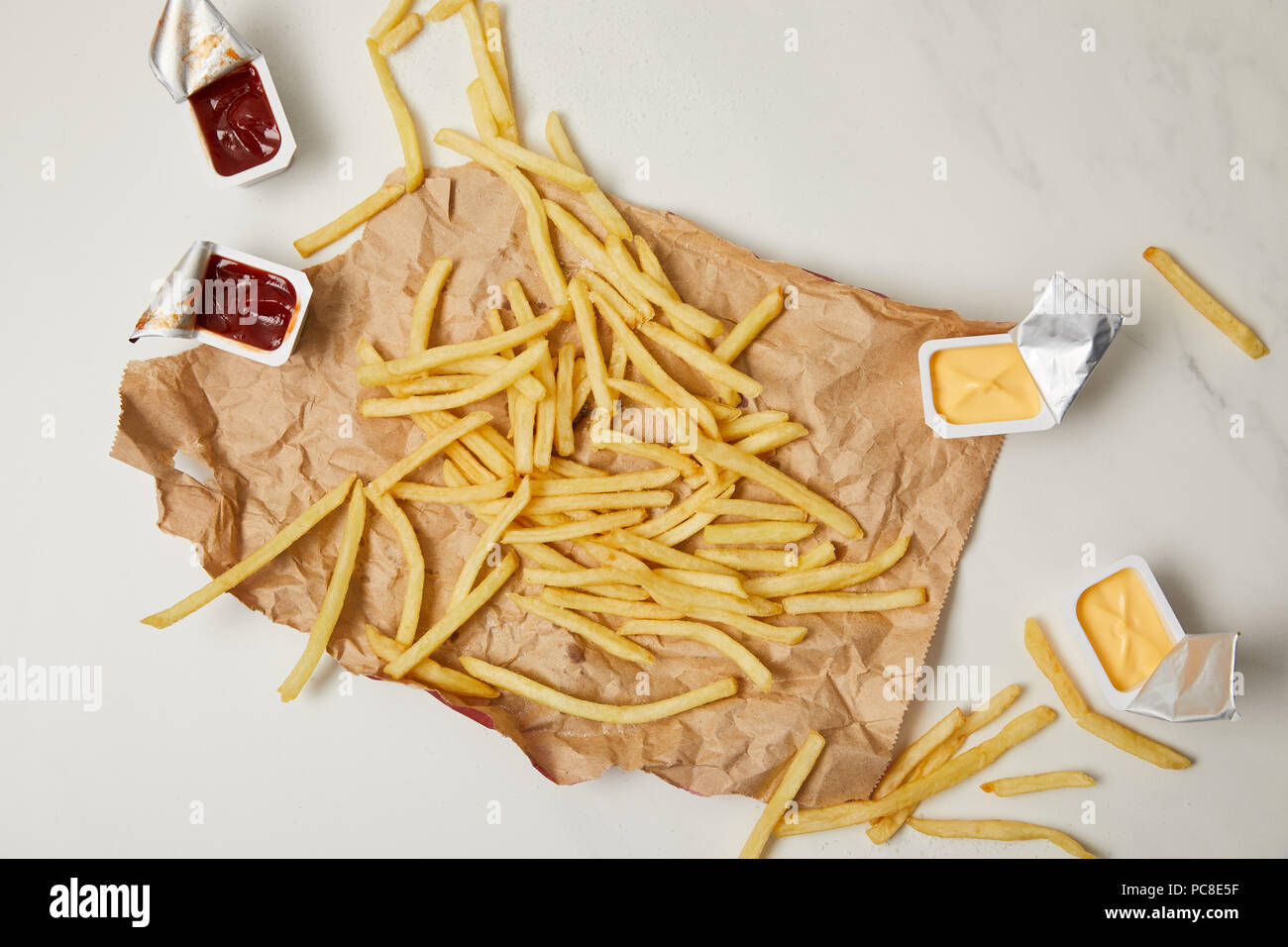 Blick von oben auf die Pommes frites auf zerknittertes Papier mit Behältern von Saucen auf Weiß Stockfoto