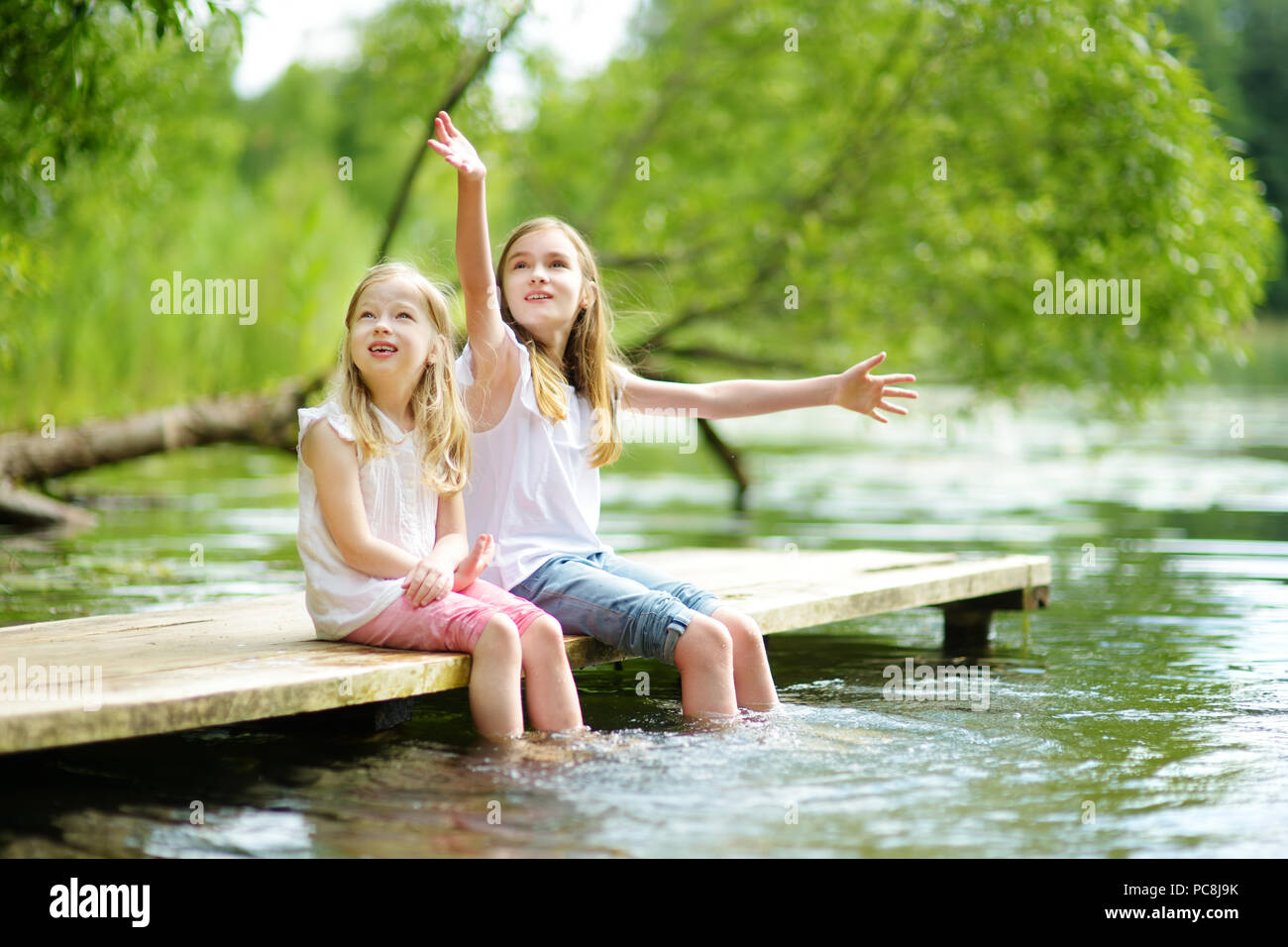 Zwei süße kleine Mädchen sitzen auf einer hölzernen Plattform durch den Fluss oder See Ihre Füße eintauchen in das Wasser an warmen Sommertagen. Aktivitäten für die ganze Familie in Summe Stockfoto