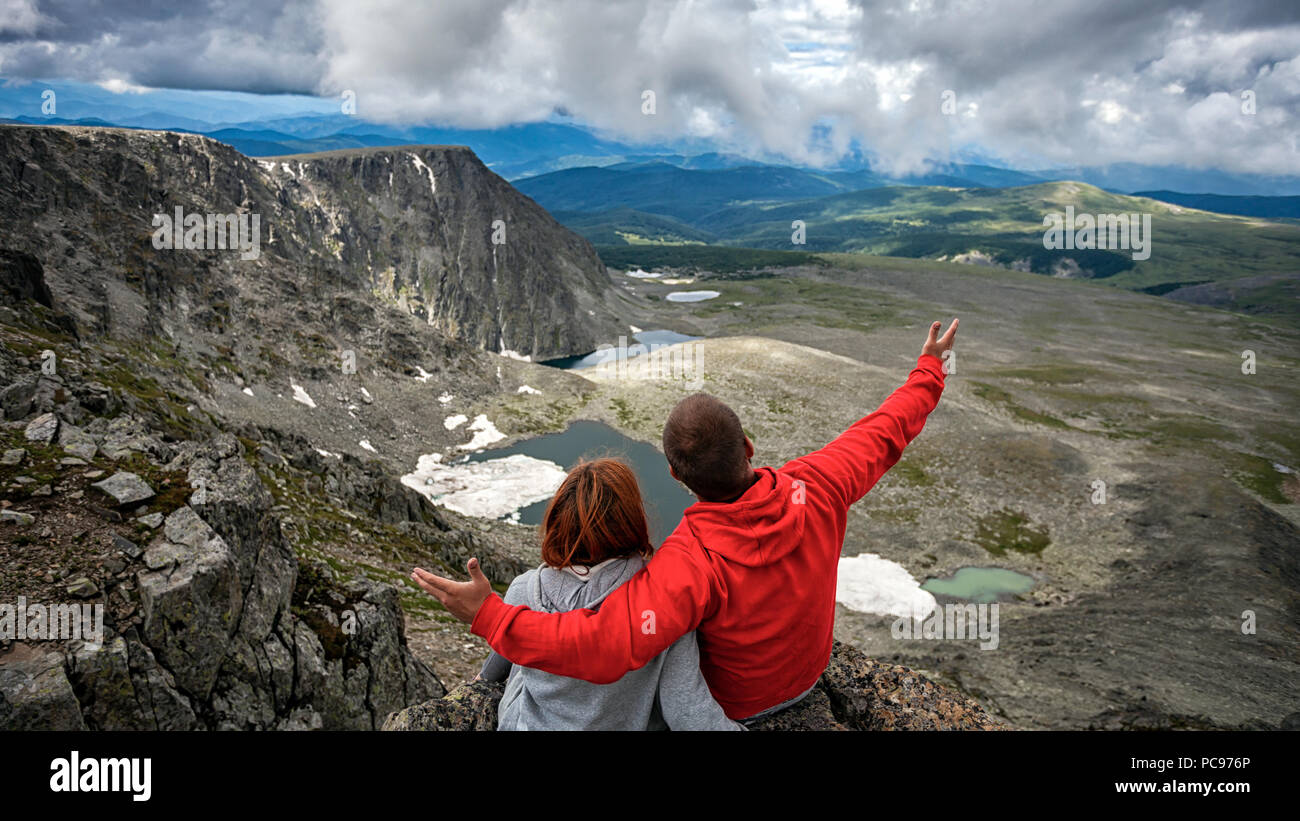 Atmosphärische moment für Liebhaber in den Bergen wandern Frau und Mann mit Rucksack sitzt auf der Oberseite der Halterung und mit Blick auf eine schöne Lan Stockfoto