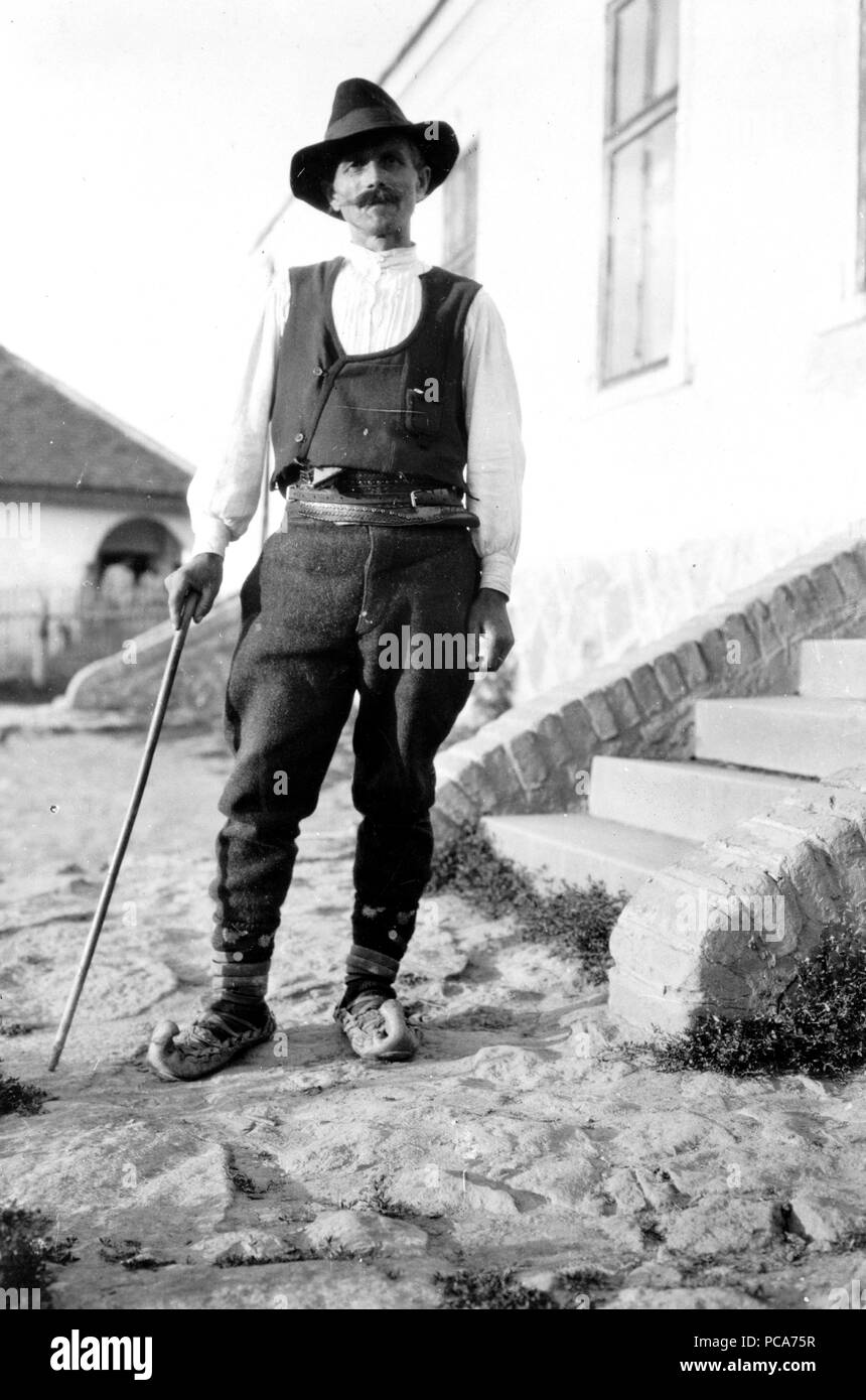 Serbische Mann mit Hut, Hemd, Hose, Gürtel und spitzen Schuhe mit Zuckerrohr stehen außerhalb der Gebäude in Jugoslawien c 1918-1924 Stockfoto