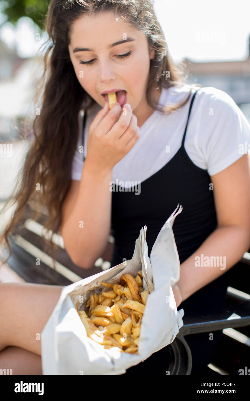 Junges Mädchen Essen Pommes Frites sitzt auf der Bank im Freien Stockfoto