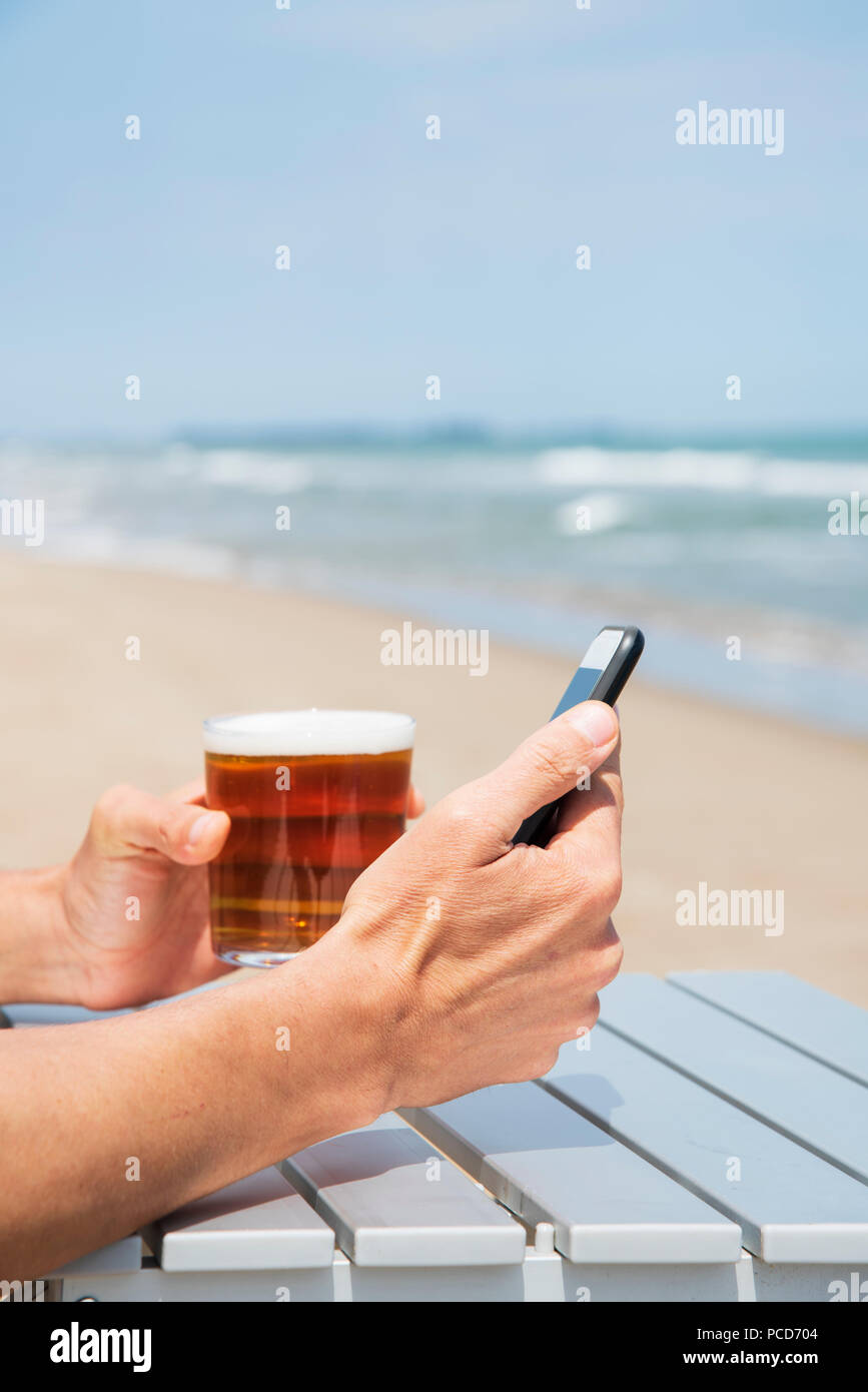 Nahaufnahme eines jungen kaukasischen Mann, an einem freien Tisch am Strand sitzend, mit einem Smartphone bei einem erfrischenden Bier Stockfoto