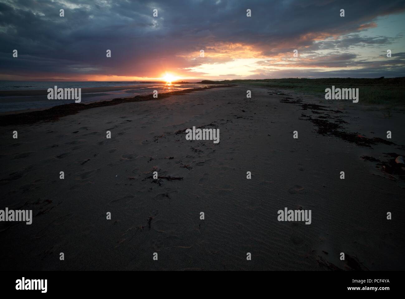 Ein Blick hinunter auf der Suche nach einem Sandstrand mit der untergehenden Sonne. Die Sonne an einem langen Sandstrand mit Seegras und Fußspuren im Sand. Stockfoto