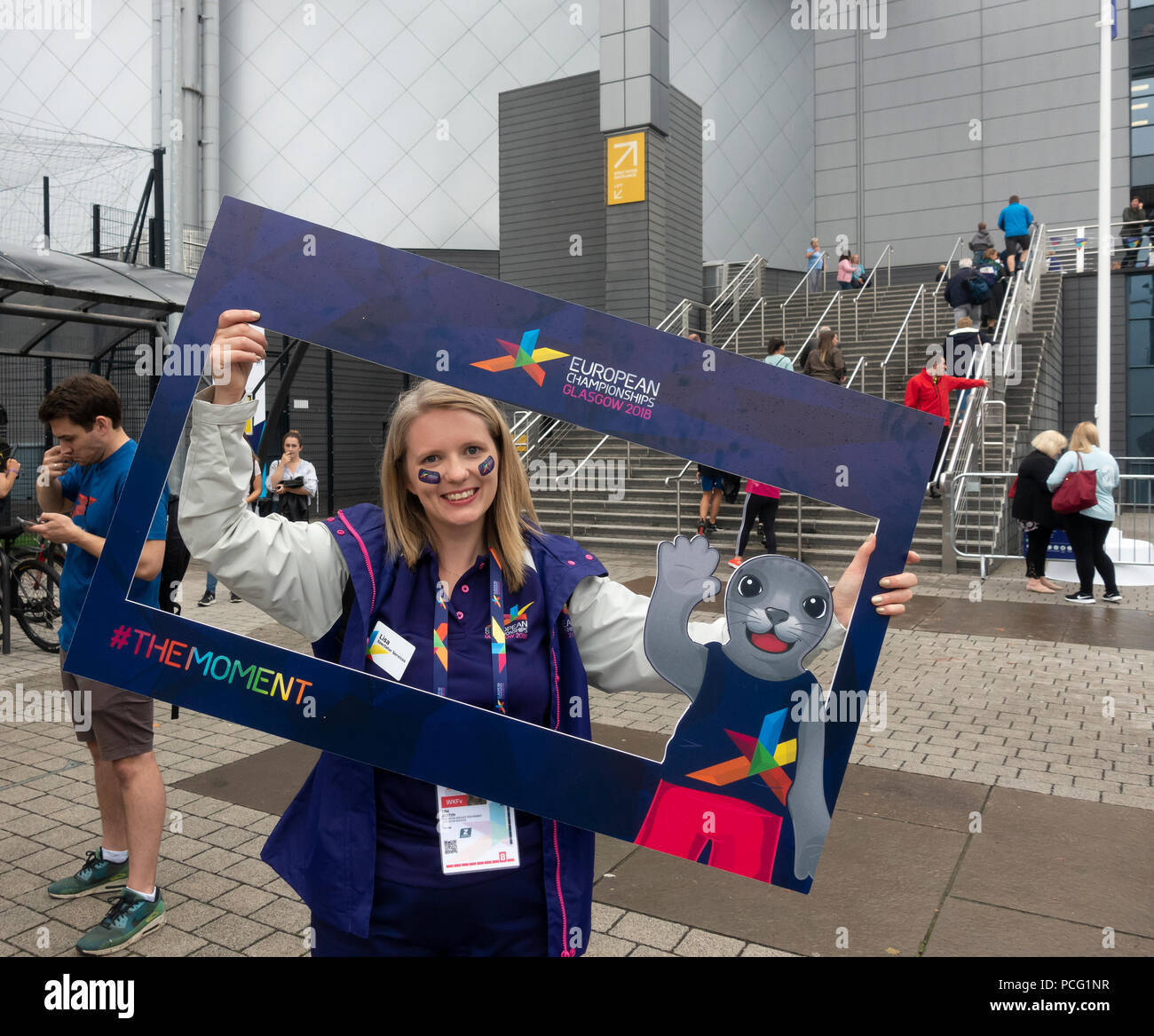 Glasgow, Schottland, Großbritannien. 02 August, 2018. Ein lächelndes Freiwillige posieren für ein Sealfie in einem Frame mit der Spiele Maskottchen Bonnie die Dichtung, außerhalb der Sir Chris Hoy Velodrome (Emirates Arena) in der Gegend von Glasgow Dalmarnock vor der ersten Veranstaltungen der Glasgow 2018, a multi-Sport Events. Die meisten Sportarten wird bewirtet, in Schottland, mit dem Velodrom Hosting die Spur Radsport. Credit: Elizabeth Leyden/Alamy leben Nachrichten Stockfoto