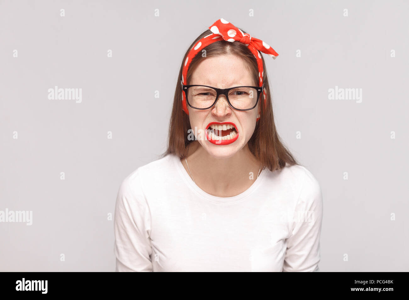 Wütend das Gesicht schreiend Portrait von Wut verrückt Bossy emotionale junge Frau im weißen T-Shirt mit Sommersprossen, schwarze Brille, rote Lippen und Kopfband. indoor Stockfoto