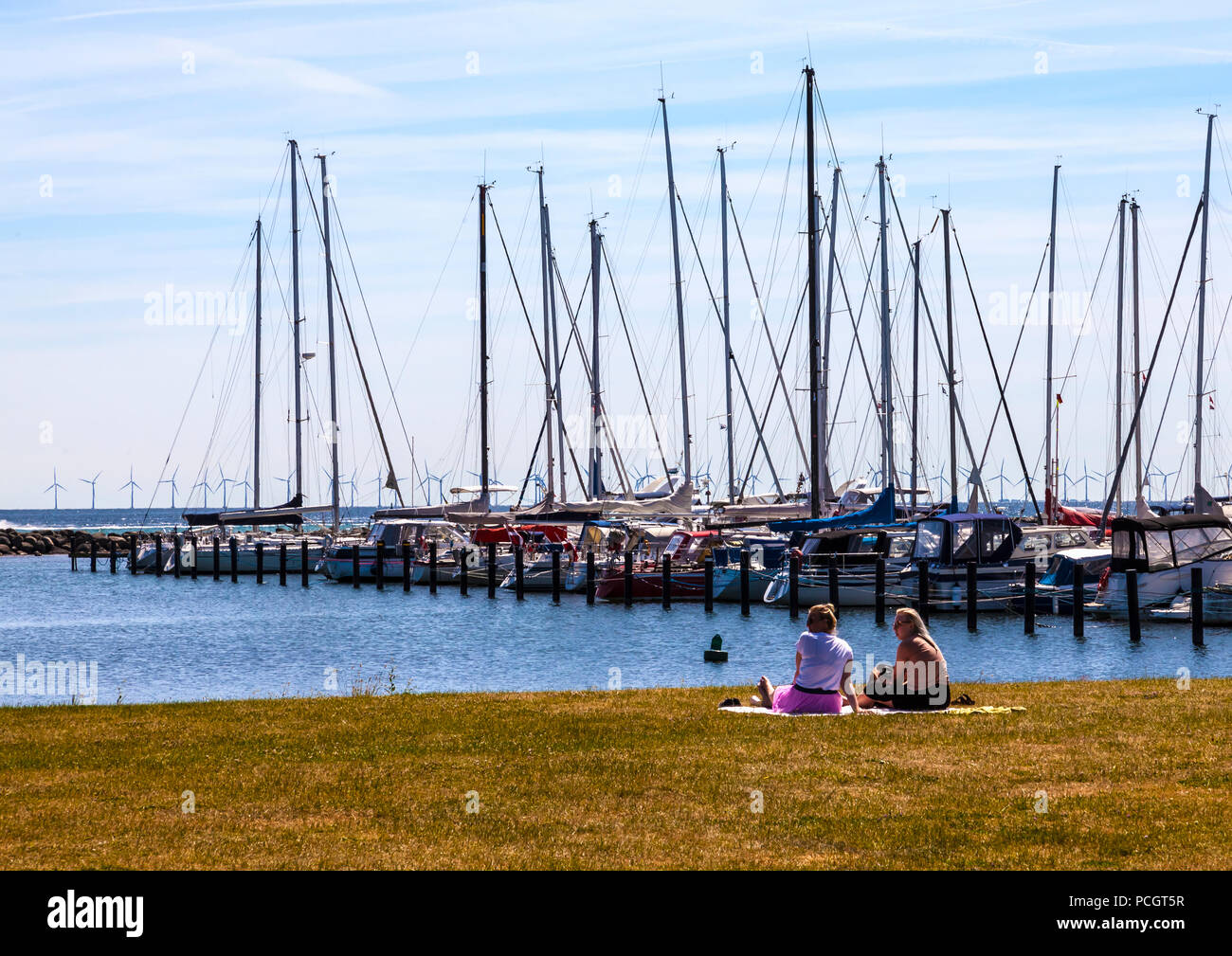 Zwei Frauen sitzen und bewundern Sie günstig Yachten in Dragør Fischerdorf in der Nähe von Kopenhagen in Dänemark mit Windparks im Hintergrund Stockfoto