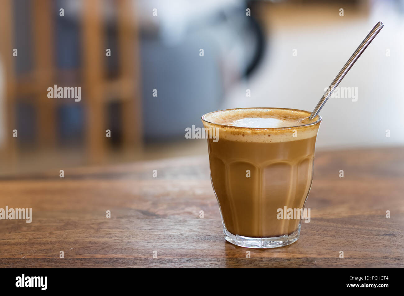 Einen heißen Kaffee mit Milch auf transparentem Glas mit einem Löffel im Inneren auf einer hölzernen Tisch, Nahaufnahme. Stockfoto