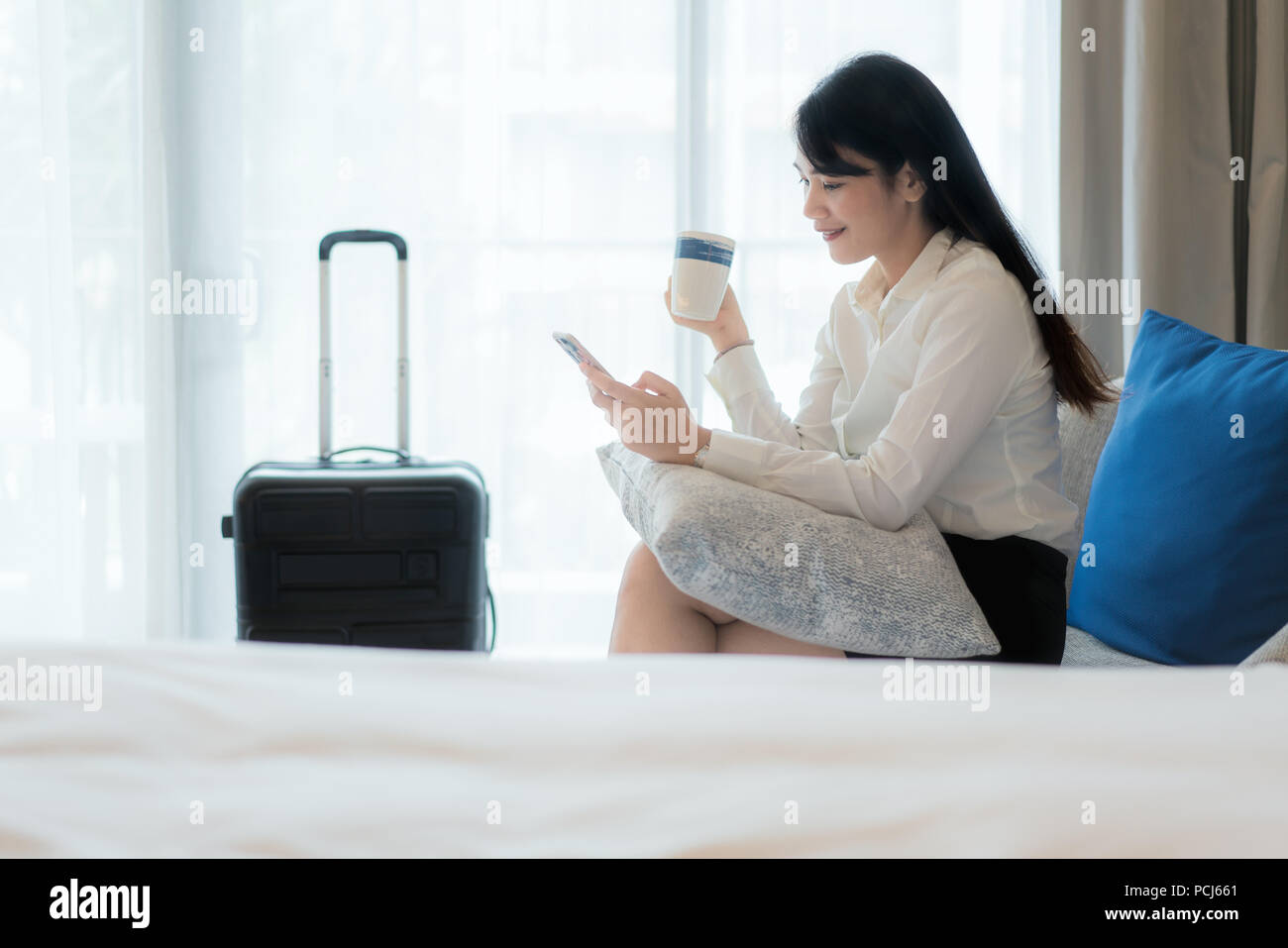 Schöne Asiatische junge lächelnde Geschäftsfrau im Anzug Kaffee trinken und über Handy beim Sitzen auf dem Sofa im Zimmer. Business Travel. Stockfoto