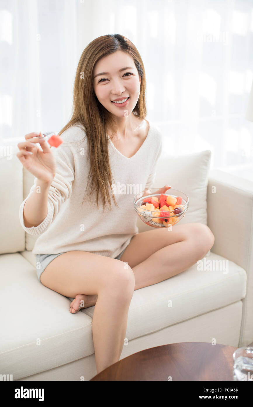 Fröhlicher junger chinesische Frau essen Obst Salat auf Sofa Stockfoto