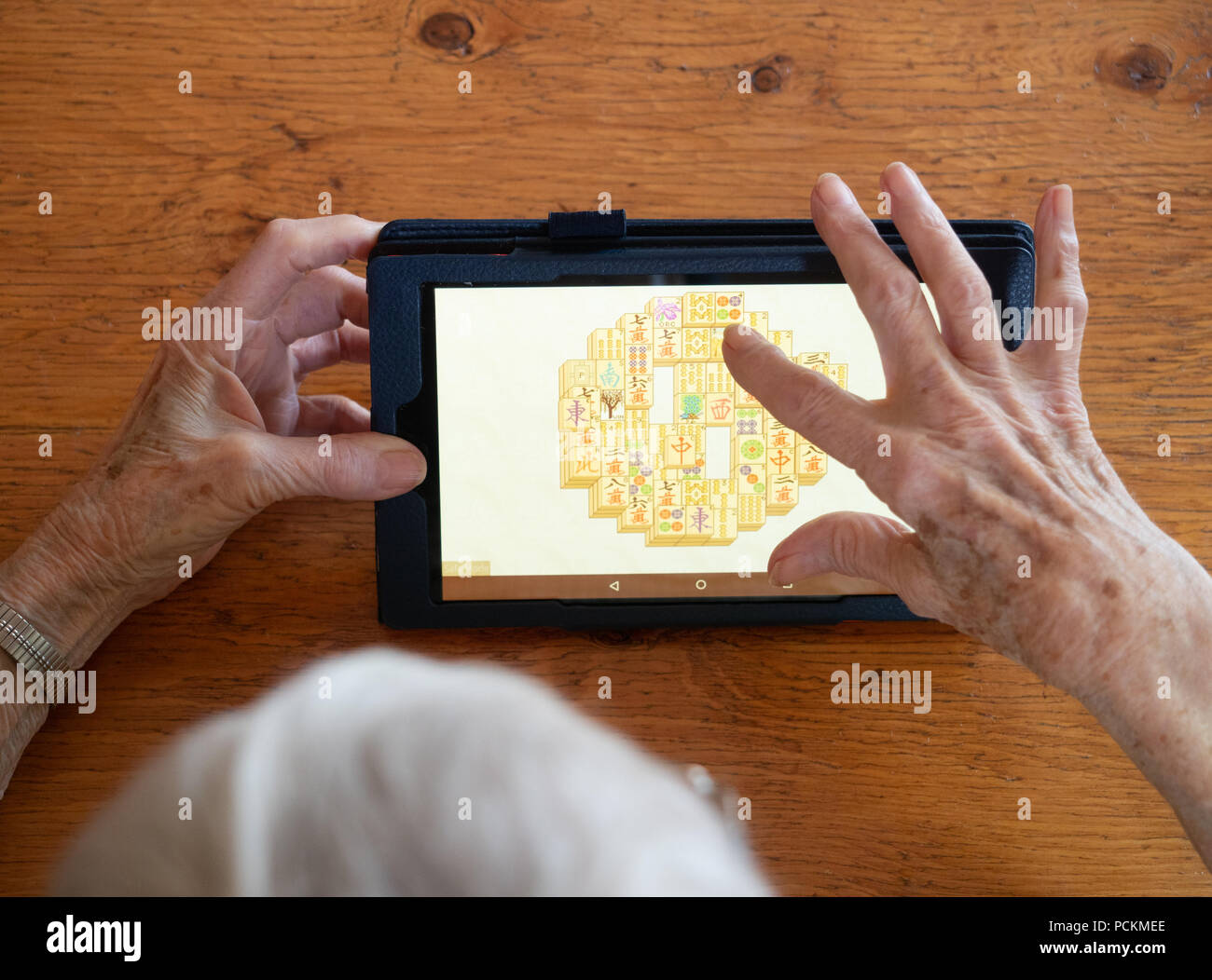 Eine ältere Frau Mahjong spielen auf einem Tablet-PC, der ein Holztisch. Nur ihre Hände und Kopf zeigen. Stockfoto