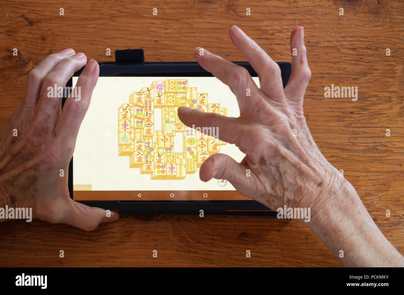 Eine ältere Frau Mahjongg spielen auf einem Tablet-PC, der ein Holztisch. Nur ihre Hände zeigen. Stockfoto
