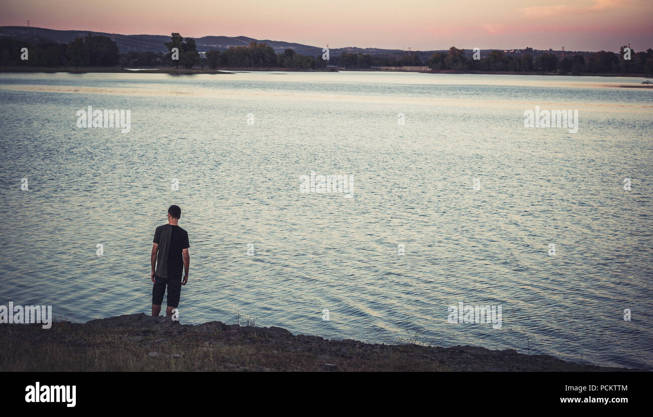 Junge Mann stand und zurück mit legere Kleidung am Ufer des Sees wieder bei Sonnenuntergang, mit Eukalyptus Wald im Hintergrund, Spanien Stockfoto