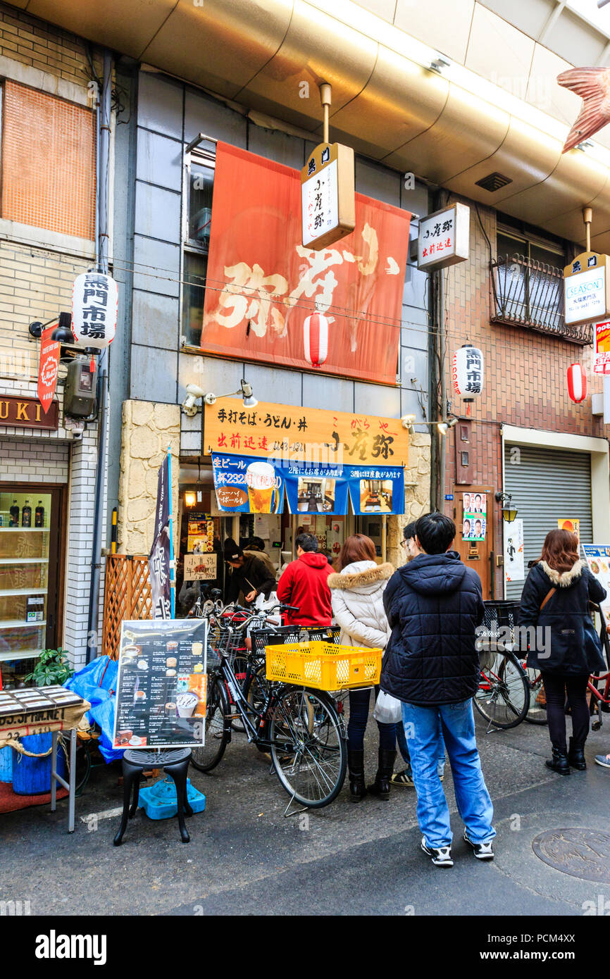 : Kuromon Ichiba, Küche essen Osaka's Markt. Die Menschen in der Warteschlange außerhalb Restaurant verkauf Soba-nudeln Buchweizen Nudeln und andere lokale Gerichte. Stockfoto