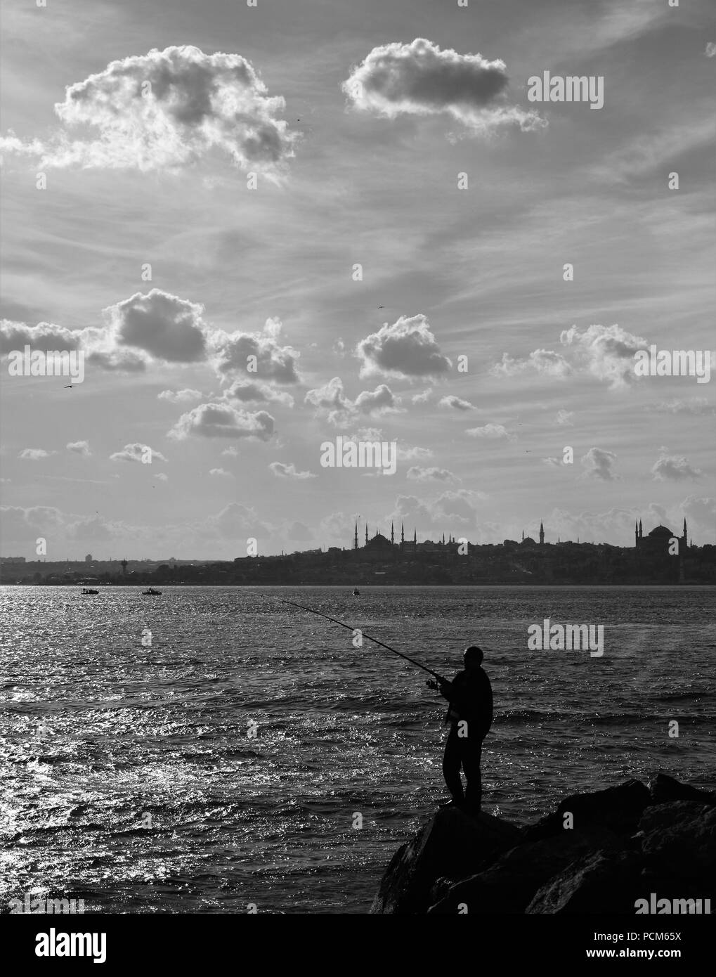 Der Mann ist die Fischerei auf dem Felsen in der Nähe des Leuchtturms und der Stadt Silhouette im Hintergrund Stockfoto