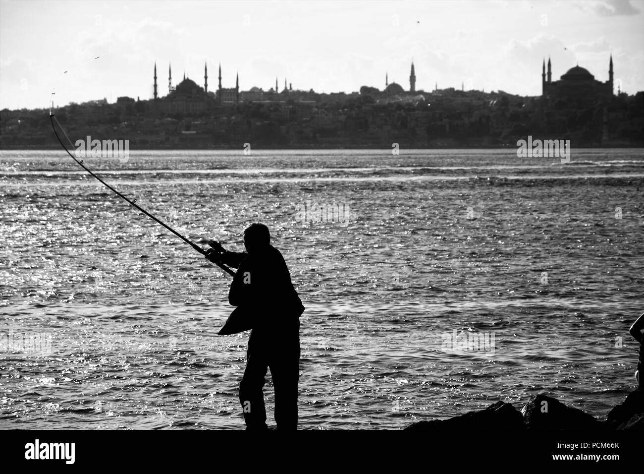 Der Mann ist die Fischerei auf dem Felsen in der Nähe des Leuchtturms und der Stadt Silhouette im Hintergrund Stockfoto