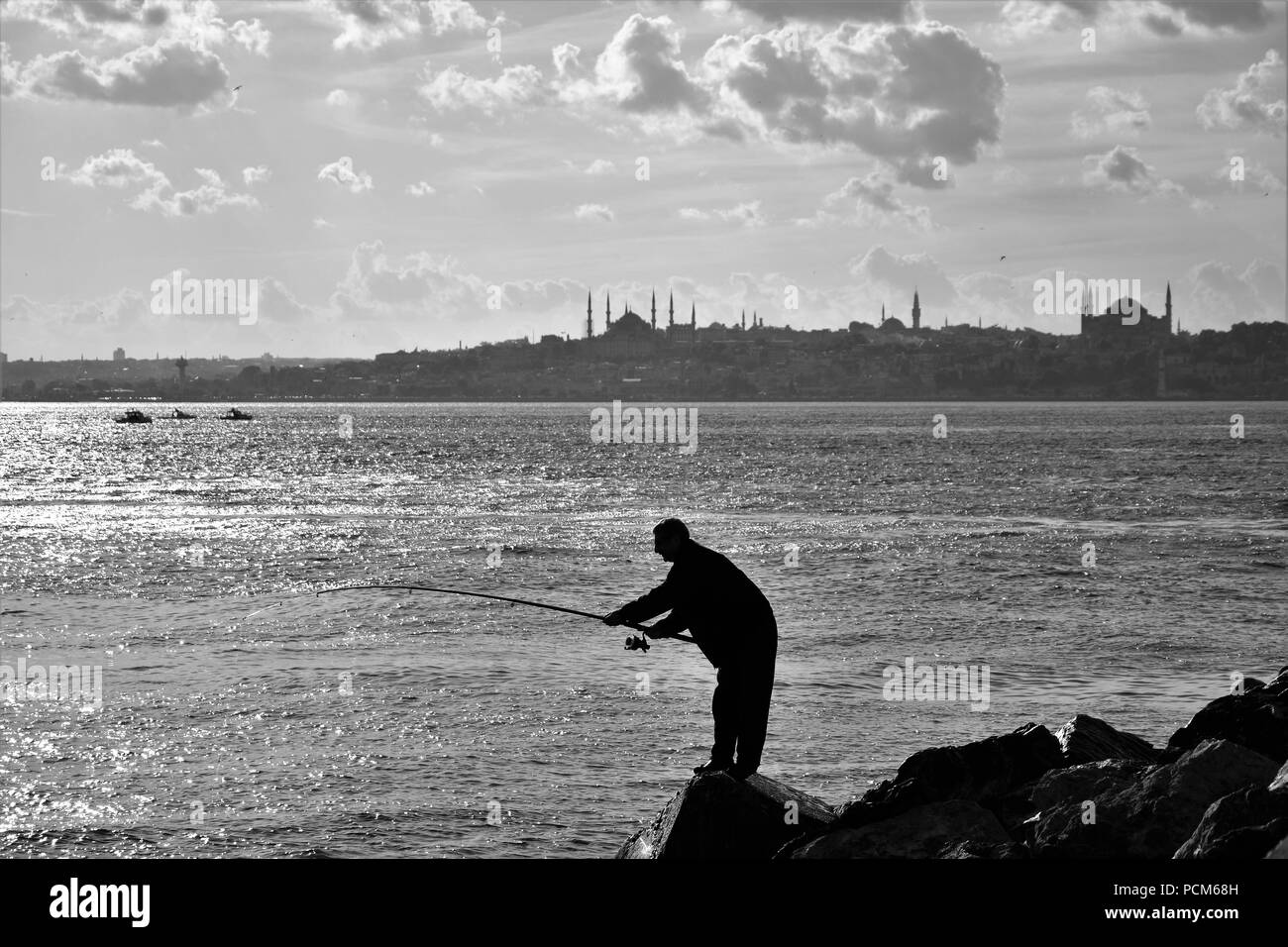 Der Mann ist die Fischerei auf dem Felsen in der Nähe des Leuchtturms und der Stadt Silhouette im Hintergrund Stockfoto