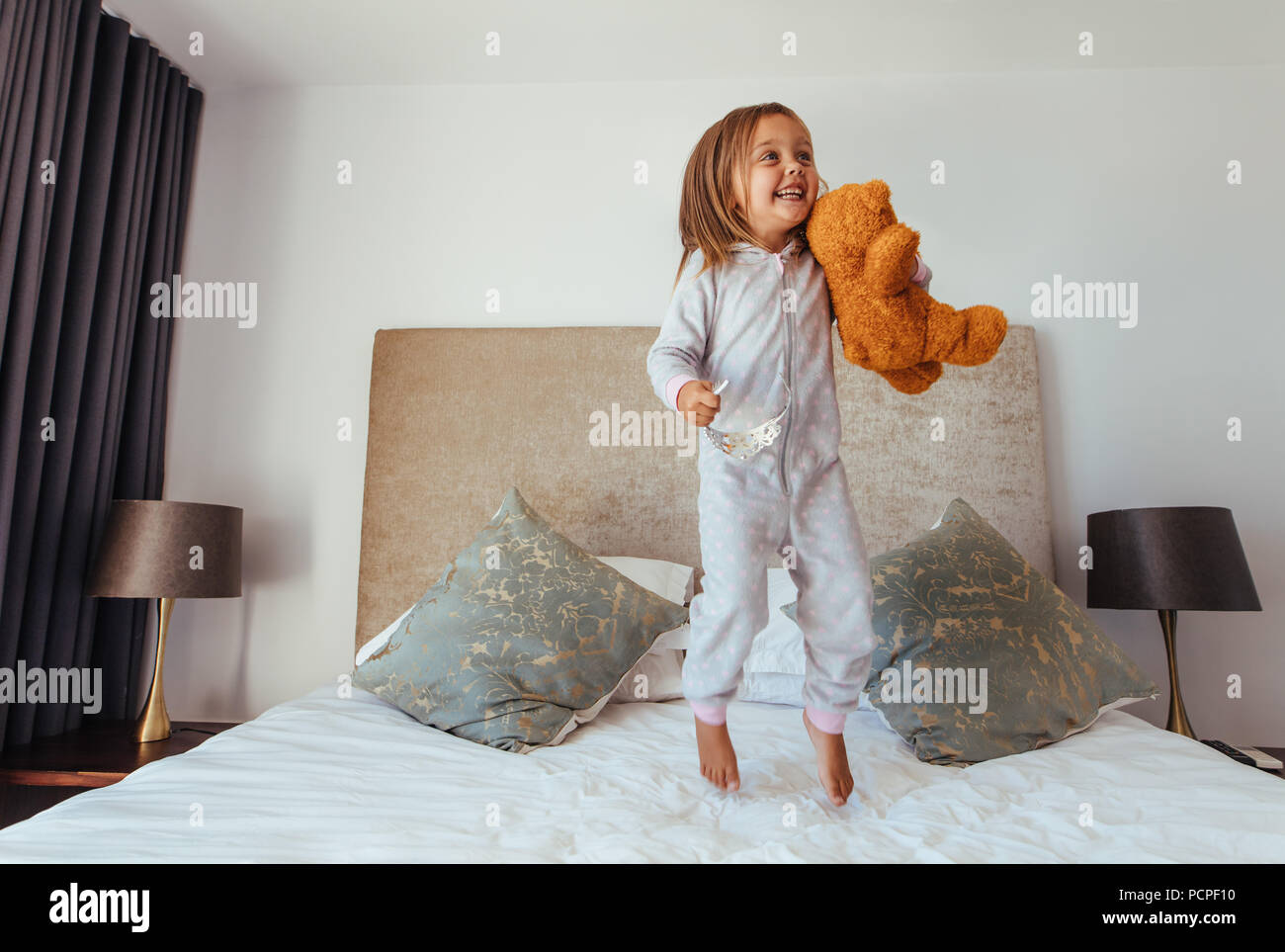 Fröhliches kleines Mädchen springen auf Bett mit Teddybär. Kind, Mädchen spielen auf dem Bett in Ihrem Schlafzimmer und lächelnd. Stockfoto