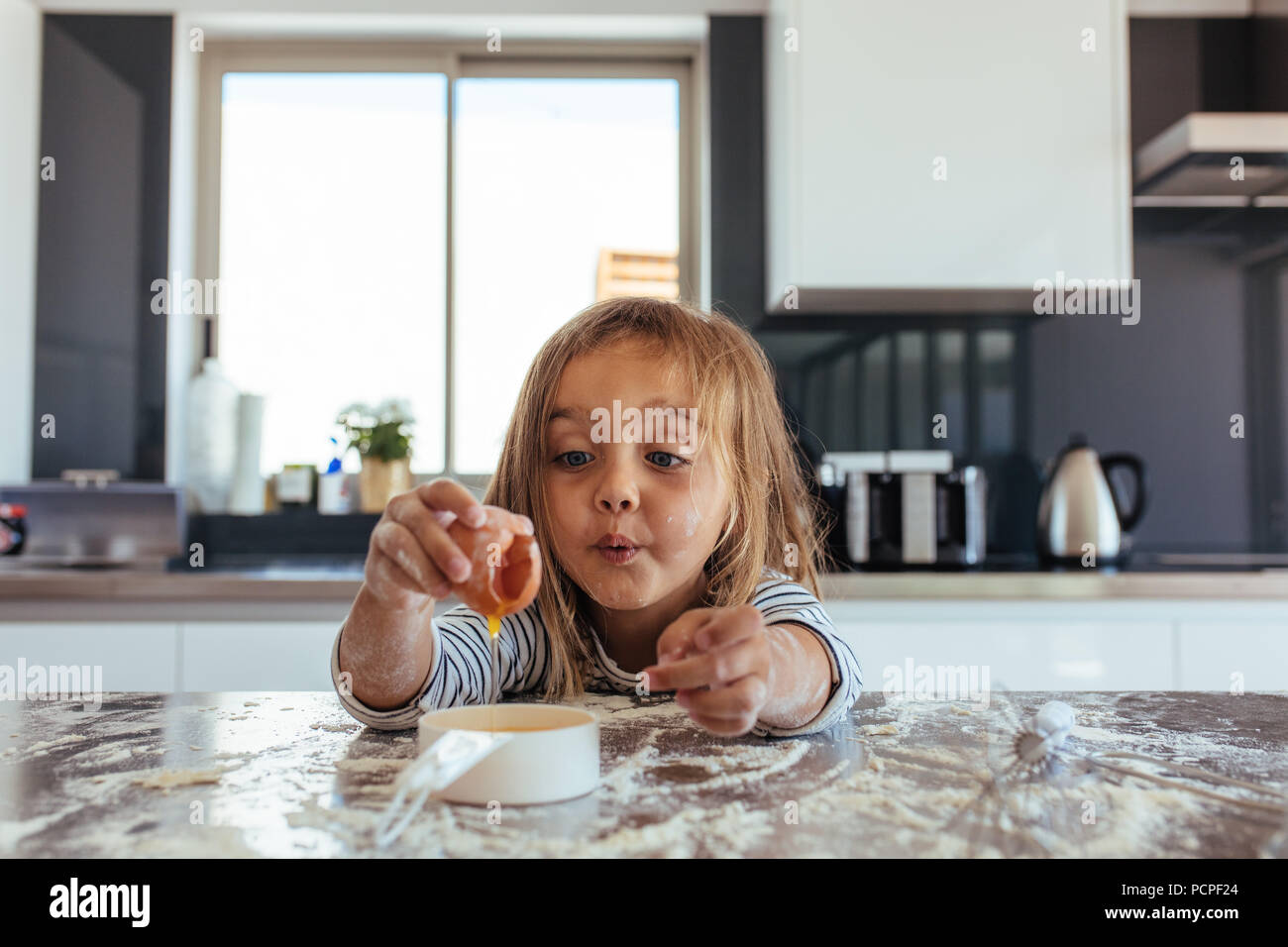 Süße kleine Mädchen brechen ein Ei in einen kleinen Schwenk über den Küchentisch. Schöne junge Mädchen backen in der Küche. Stockfoto