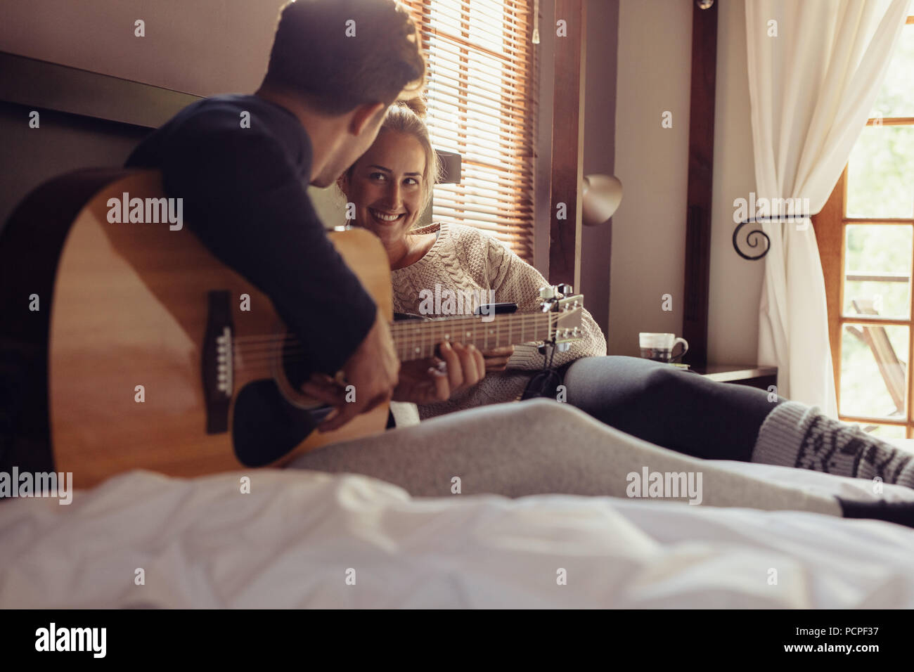 Junger Mann mit Gitarre für seine Freundin im Bett. Paar Ruhe im Hotel Zimmer und Gitarre spielen im Bett. Stockfoto
