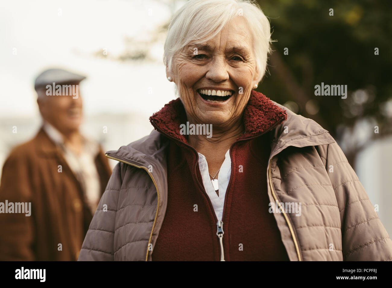 Portrait von fröhlichen älteren Frau in warme Kleidung an Kamera und lachend mit einem Mann im Hintergrund. Alte Frau im Freien an einem Wintertag. Stockfoto