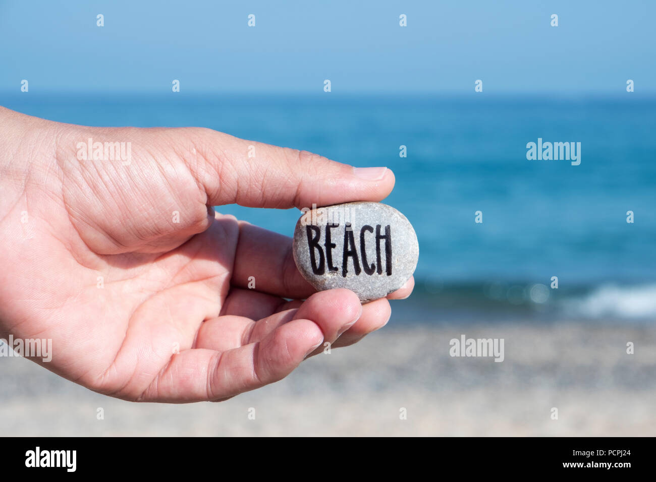 Nahaufnahme der Hand eines jungen kaukasischen Mann am Strand, vor dem Ozean, mit einem Stein mit dem Text Strand geschrieben Stockfoto