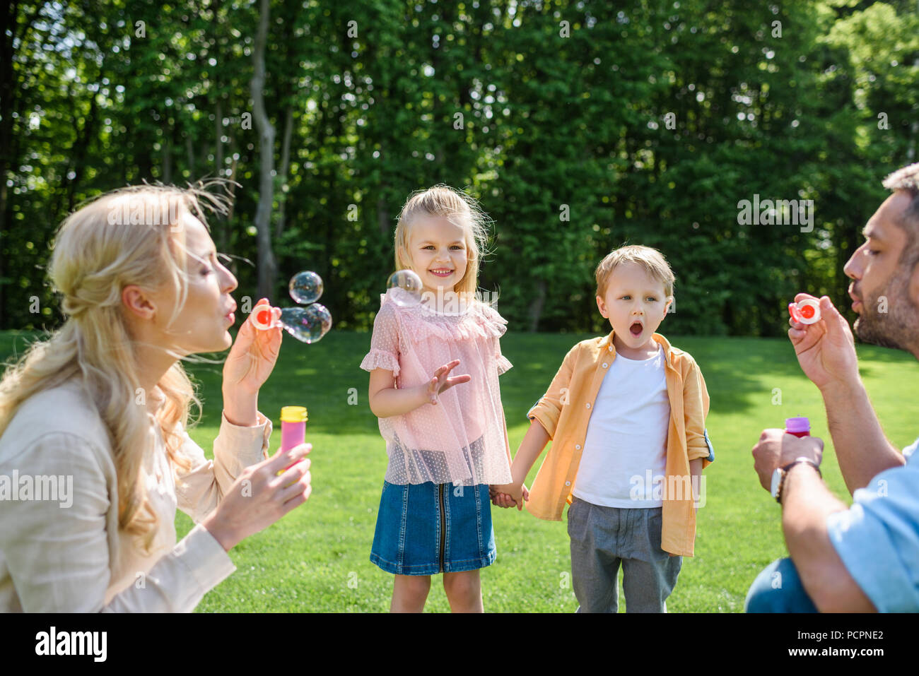 Adorable Kinder halten sich an den Händen und betrachten Eltern bläst Seifenblasen in Park Stockfoto