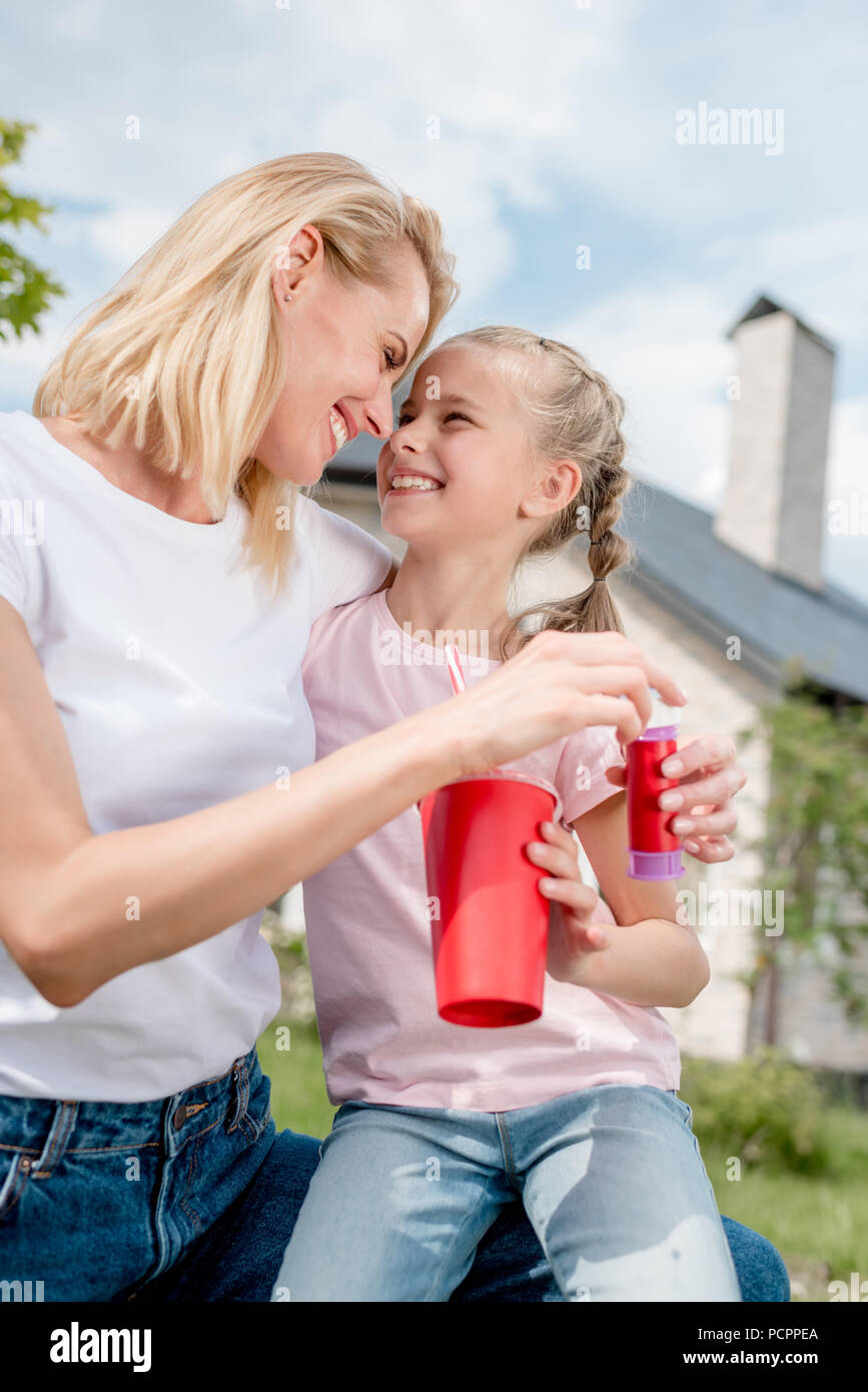 Frau mit Bubble Gebläse in der Hand sitzen, Stirn an Stirn mit Tochter holding Tasse Cola vor neuen Cottage Stockfoto